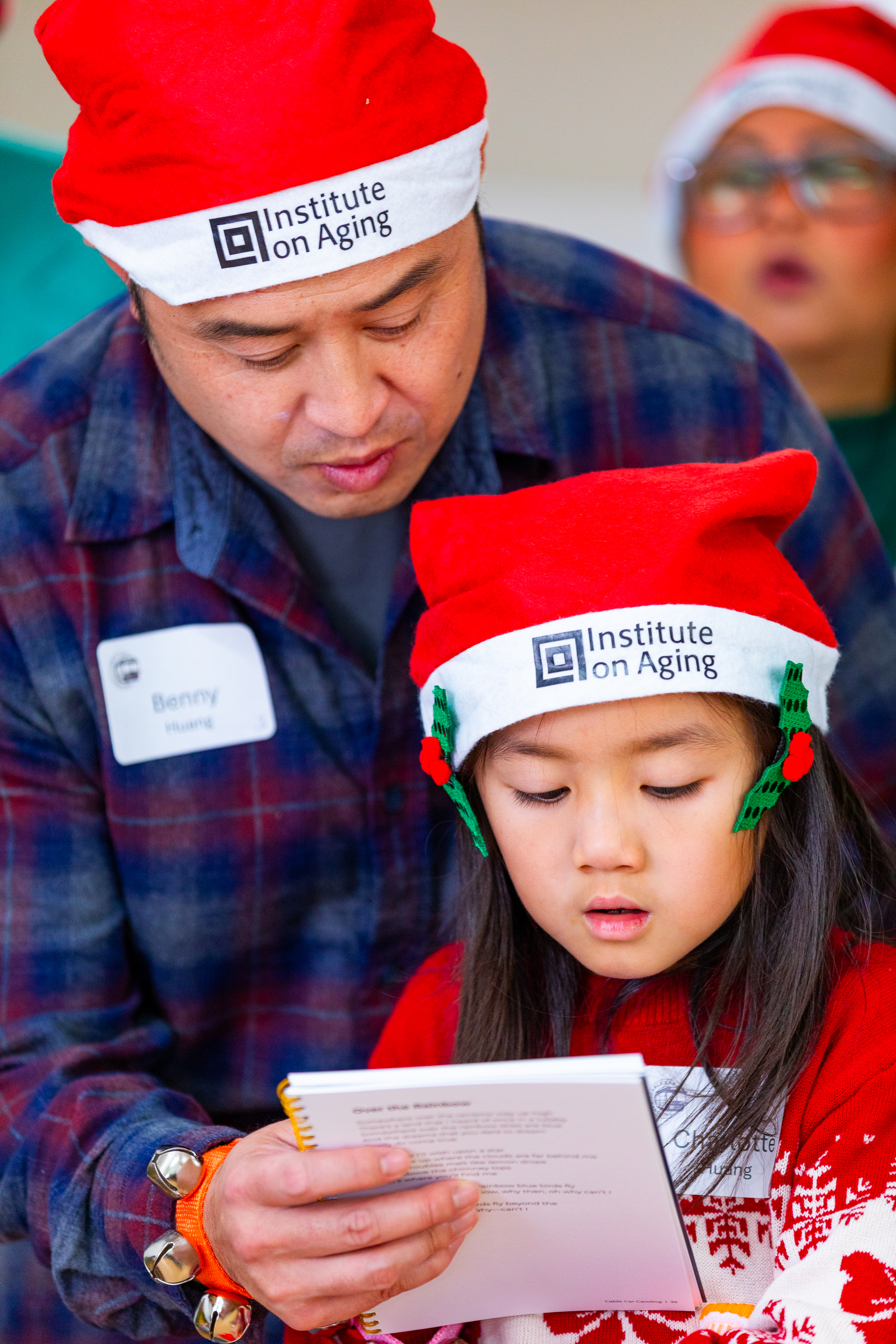 A man and a child wearing red Santa hats with &quot;Institute on Aging&quot; logos read a booklet together. The child looks focused and wears a festive red sweater.