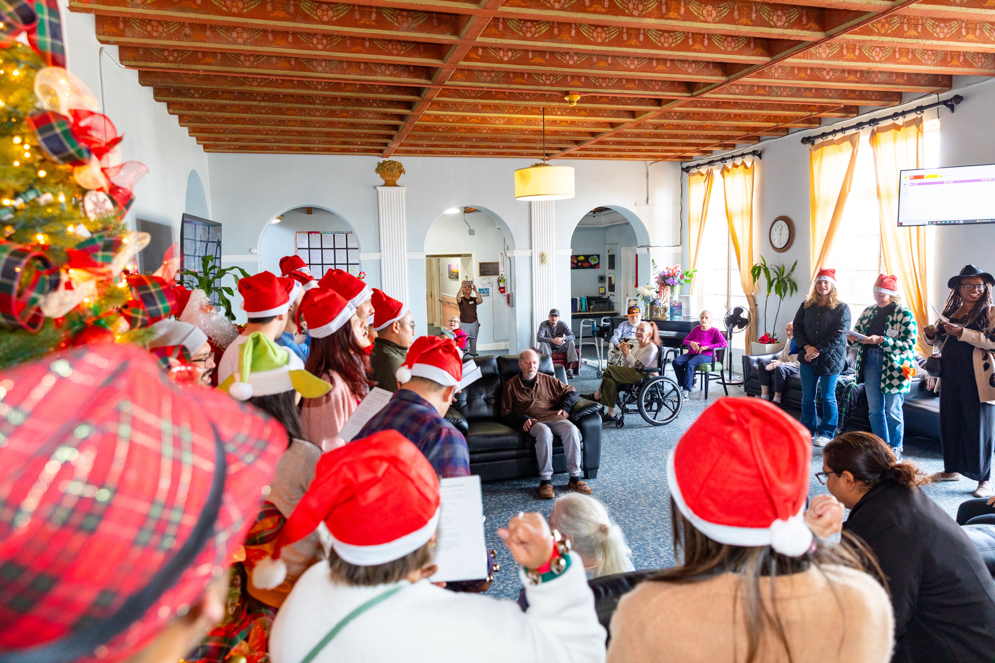 People wearing Santa hats sing to a seated audience in a festive room with Christmas decorations and a tree. The atmosphere is warm and cheerful.