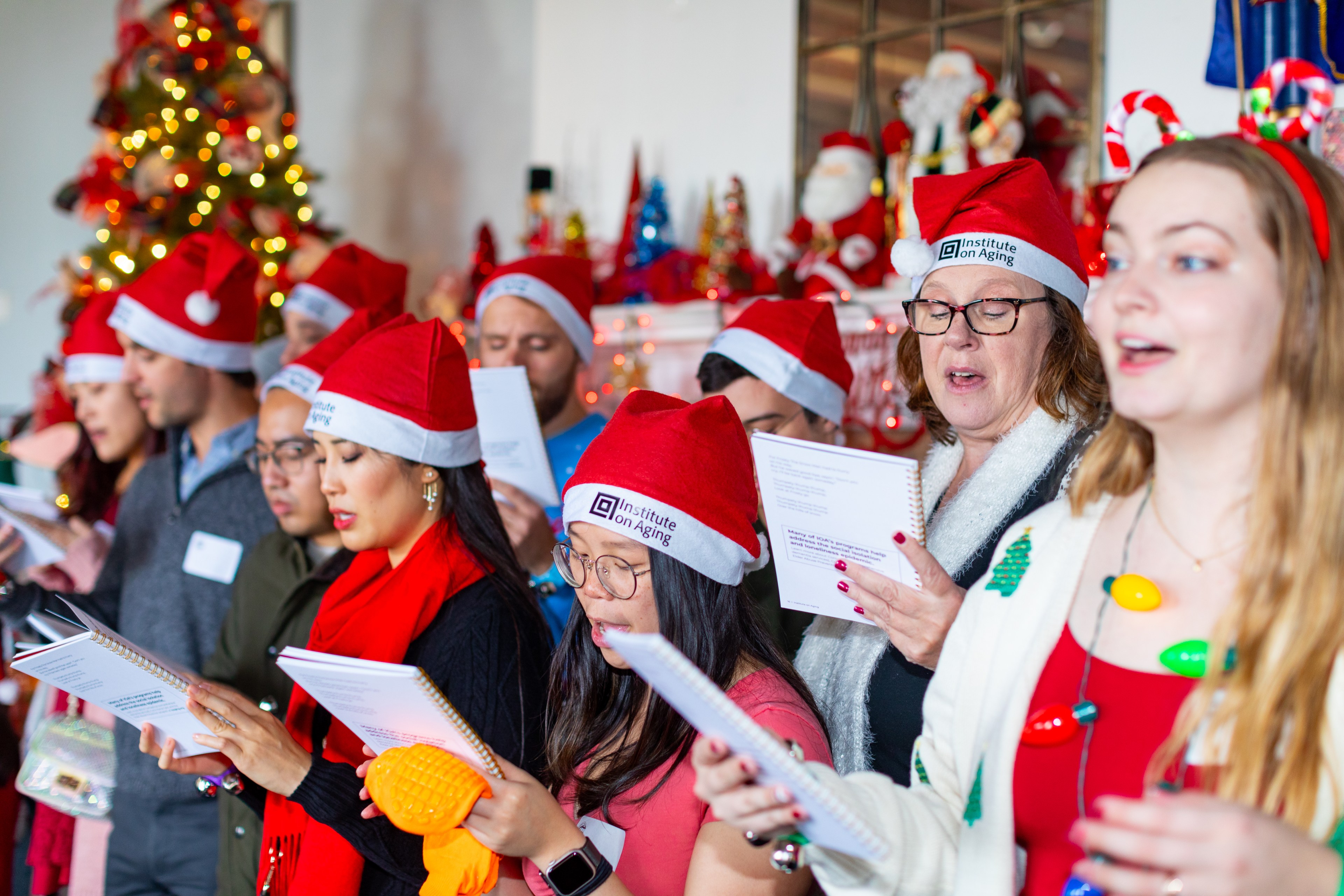 A group of people wearing Santa hats sing Christmas carols, holding songbooks, with a decorated tree and festive lights in the background.