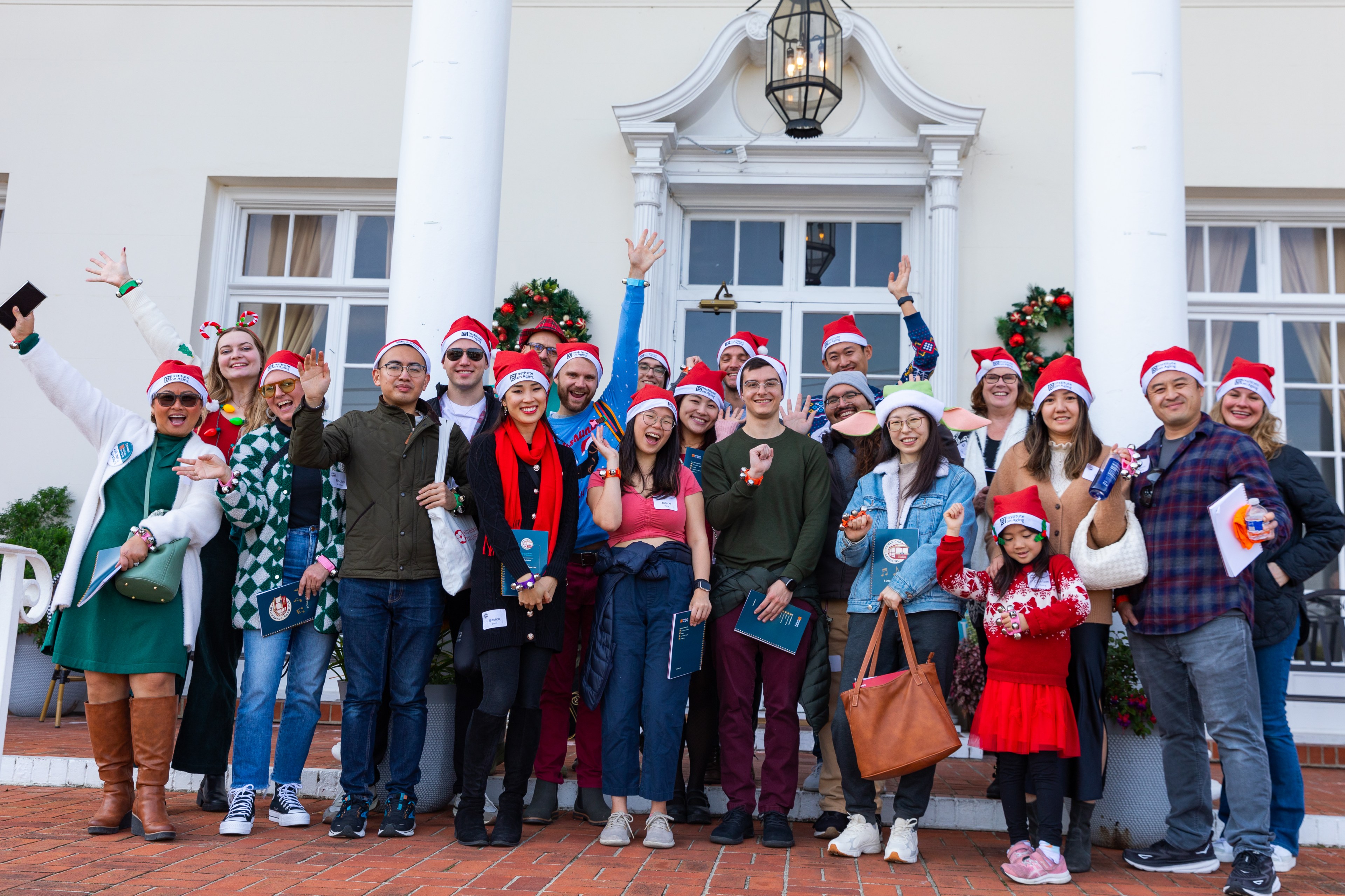 A group of people wearing Santa hats, many waving and smiling, stand in front of a festive building decorated with Christmas wreaths and lights.