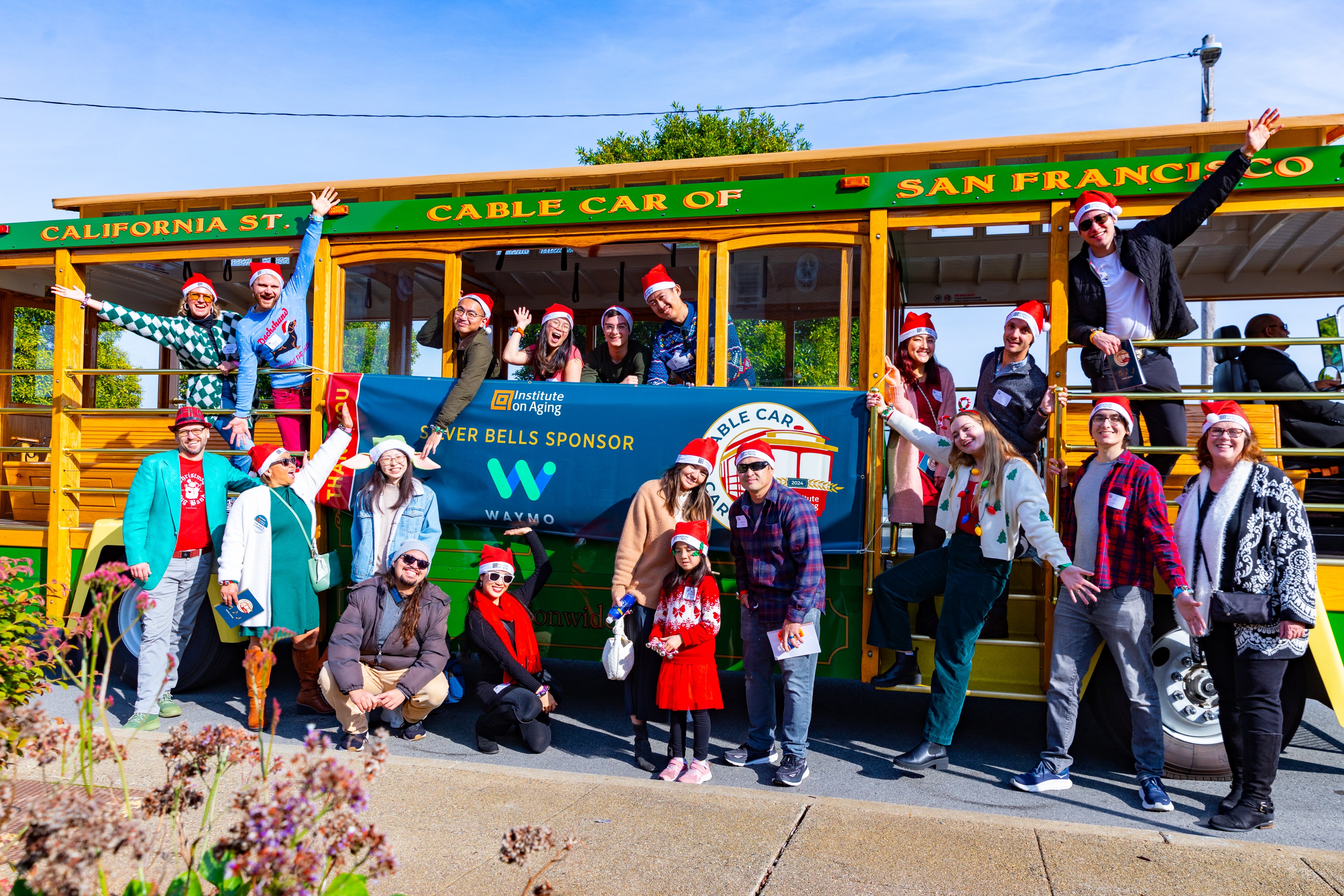 A group of people in festive attire and Santa hats pose joyfully beside a San Francisco cable car. They are standing around and inside the car, smiling brightly.