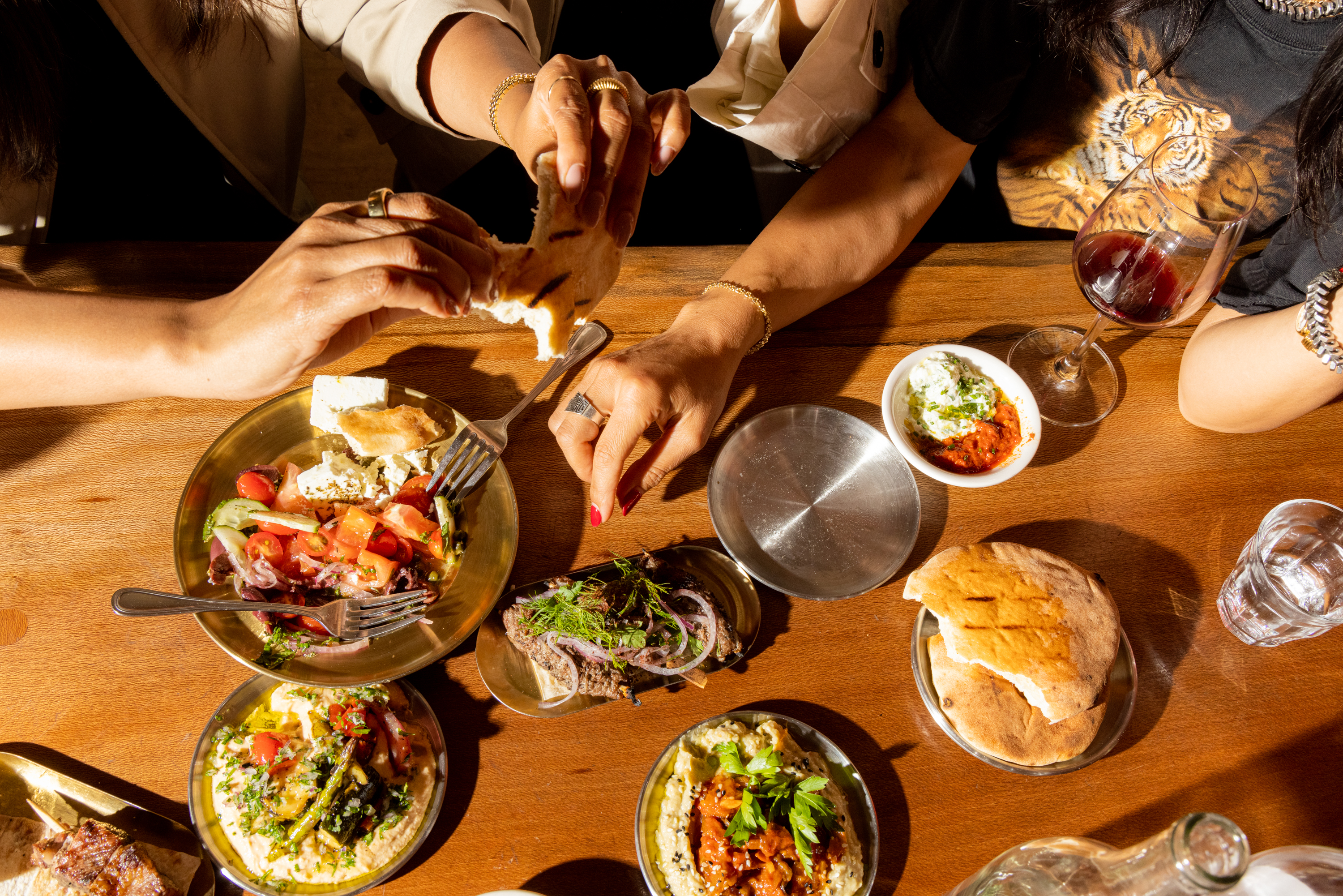 A wooden table with various dishes, including salads, dips, and grilled bread. Two people are sharing food, with glasses of wine and water nearby.