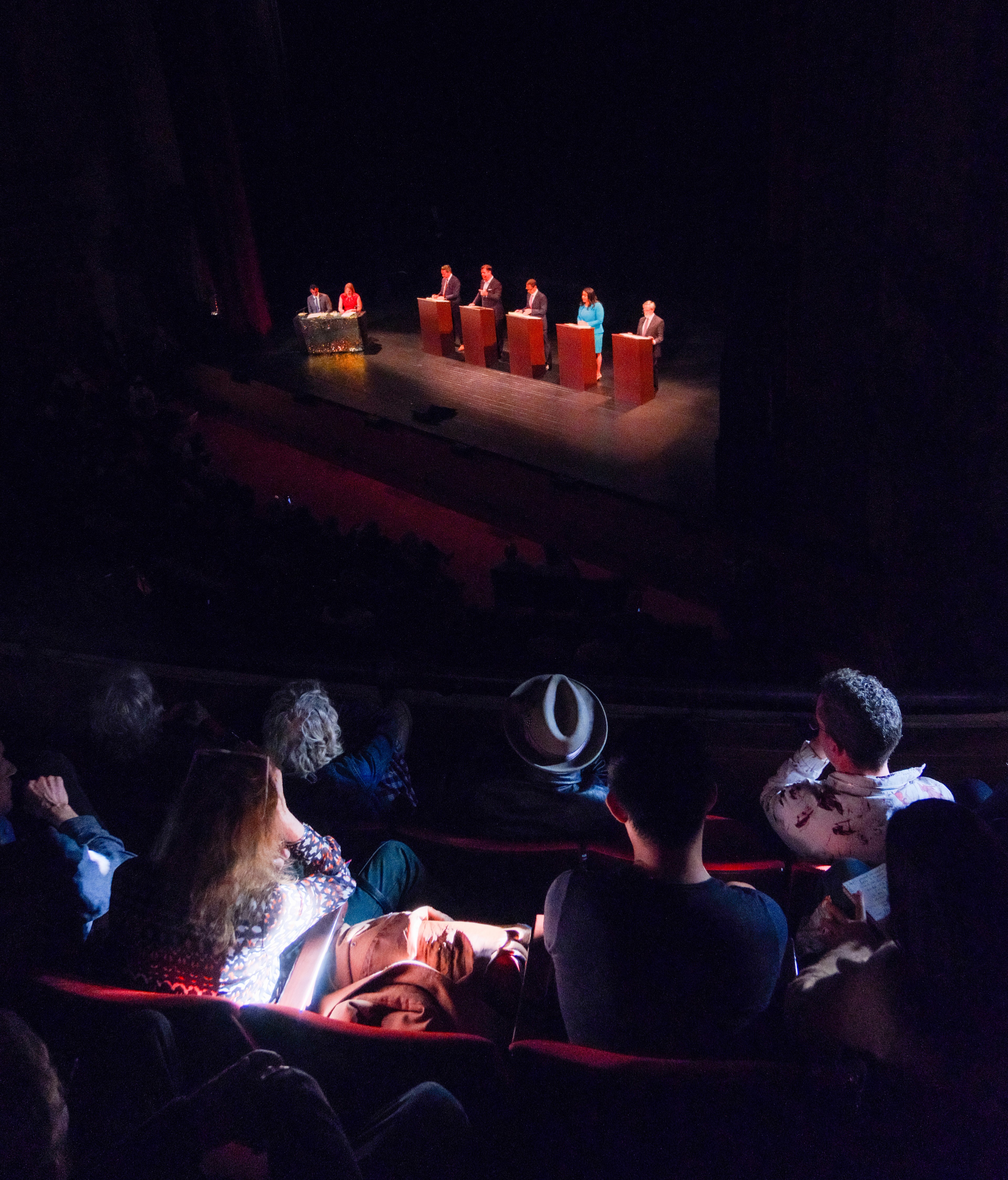 A dimly lit theater features a debate or panel discussion on stage with six people behind podiums. The audience is visible in the foreground.