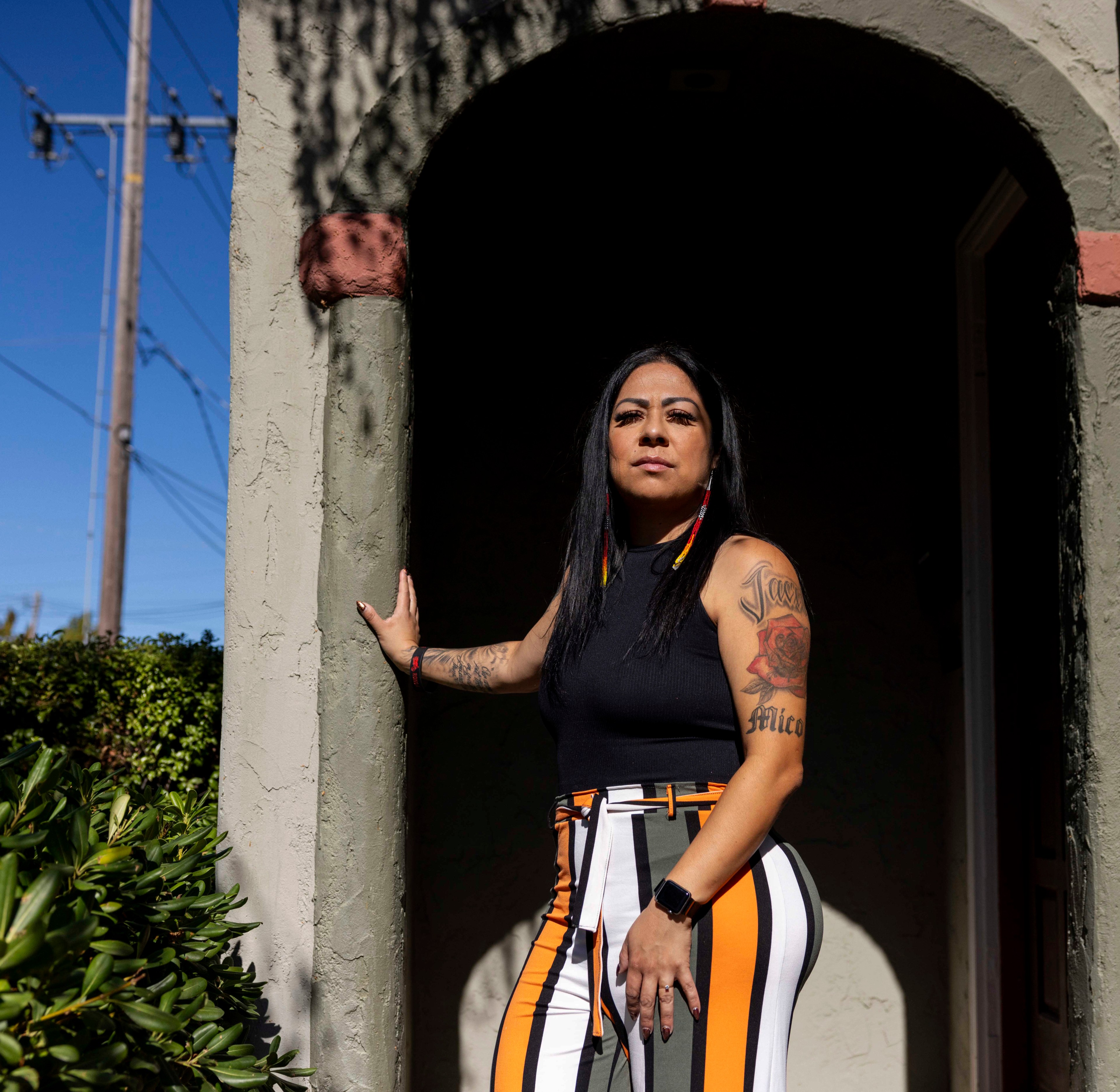 A woman stands in a sunlit archway. She has long black hair and colorful earrings. Her outfit features bold stripes, and she has tattoos on her arms.