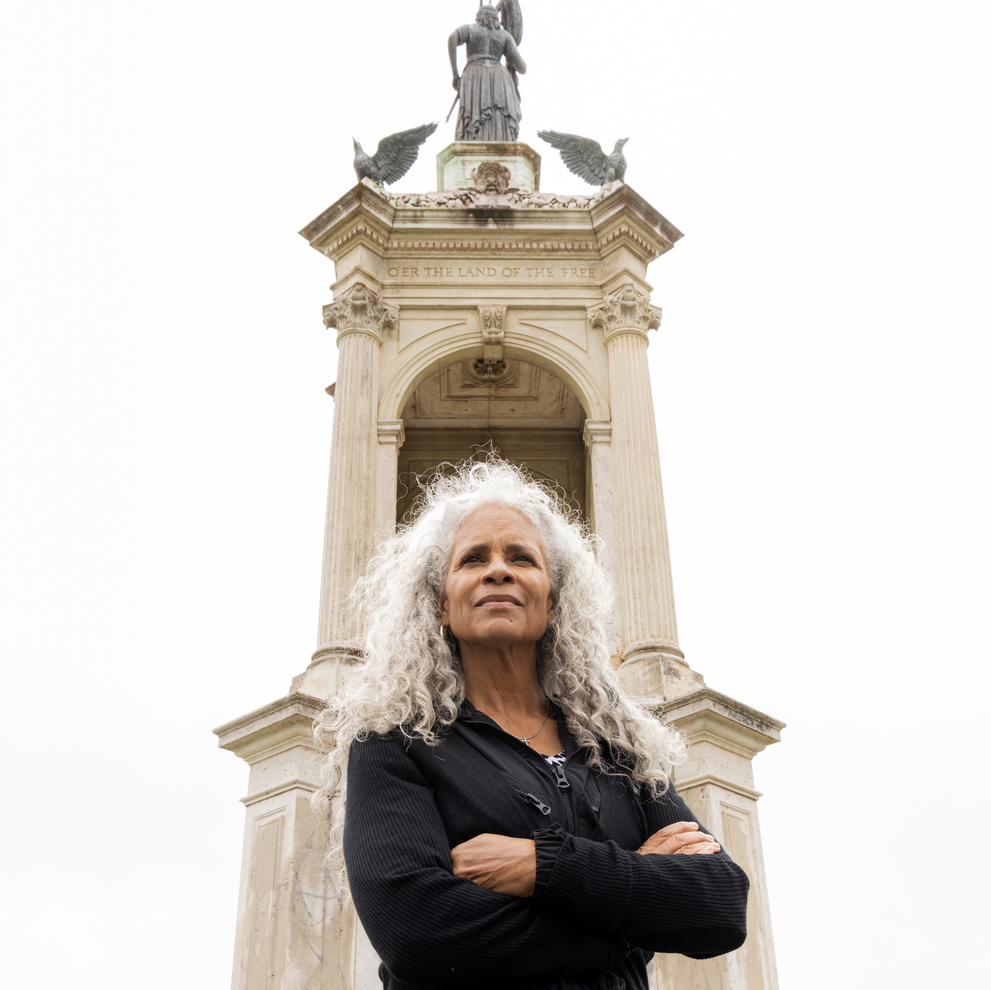 A person with long gray hair stands confidently with arms crossed, in front of a tall monument with columns and a statue on top. The sky is overcast.