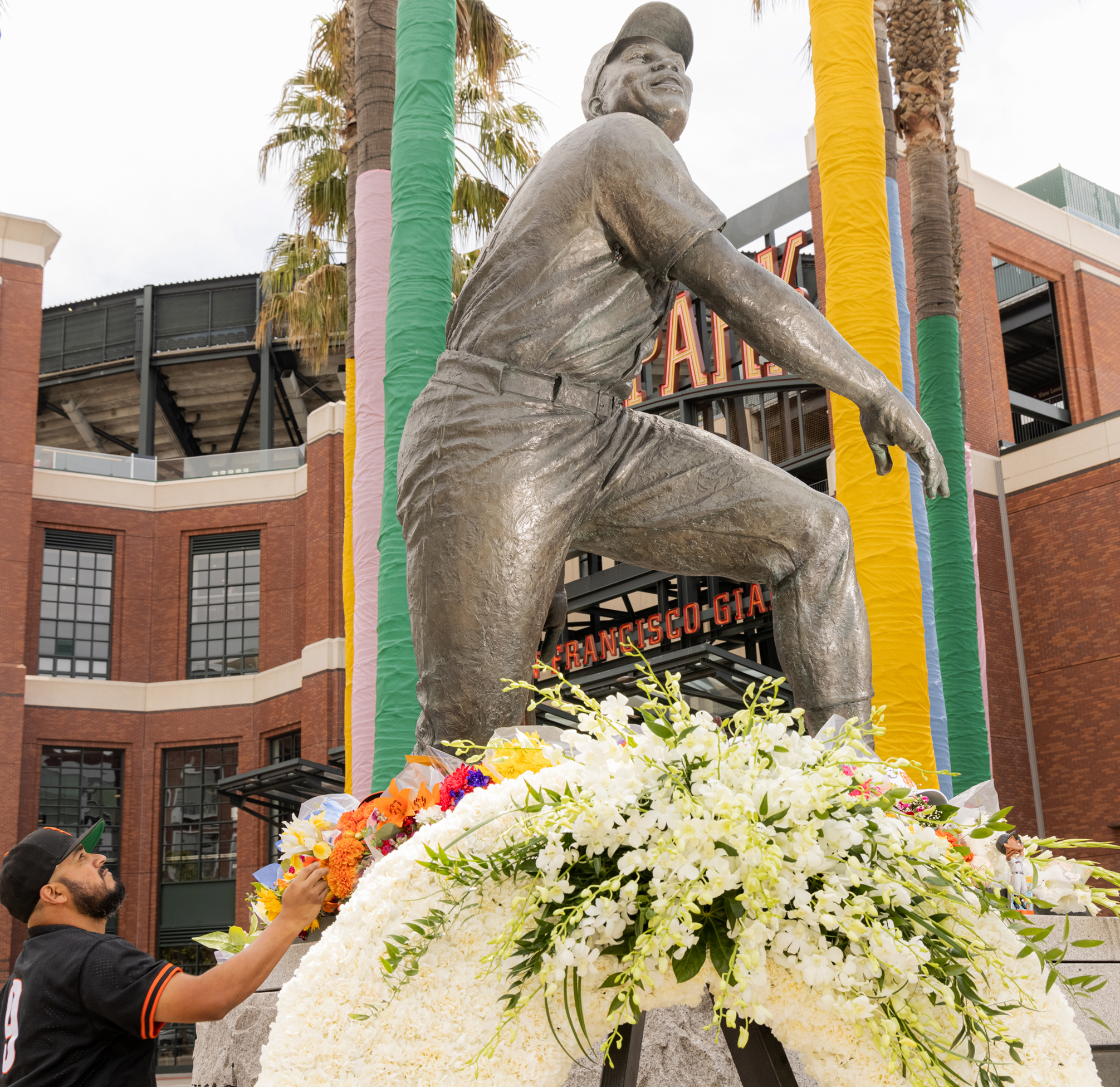 A man in a black jersey places flowers at the base of a large statue of a baseball player outside a stadium. The statue’s base is adorned with colorful flowers.