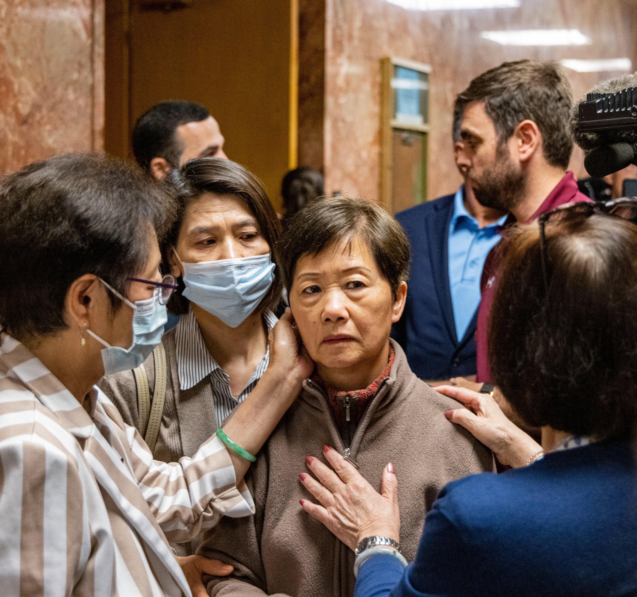 A group of people stand closely together, some with facial masks, appearing somber and supportive with gentle touches on the arm and shoulder.