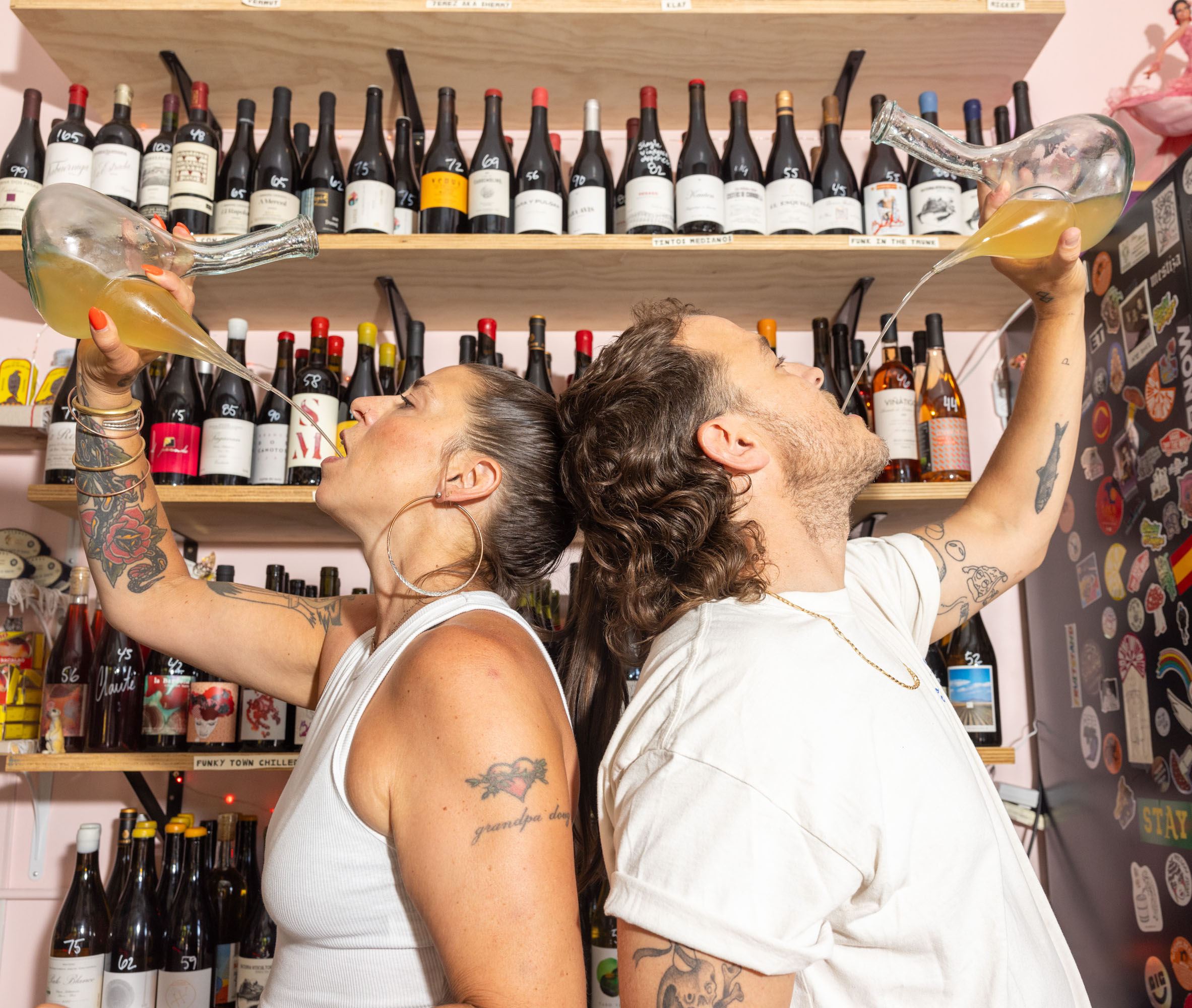 Two people are drinking from glass pitchers in front of shelves filled with wine bottles. Both have tattoos and wear casual white tops.