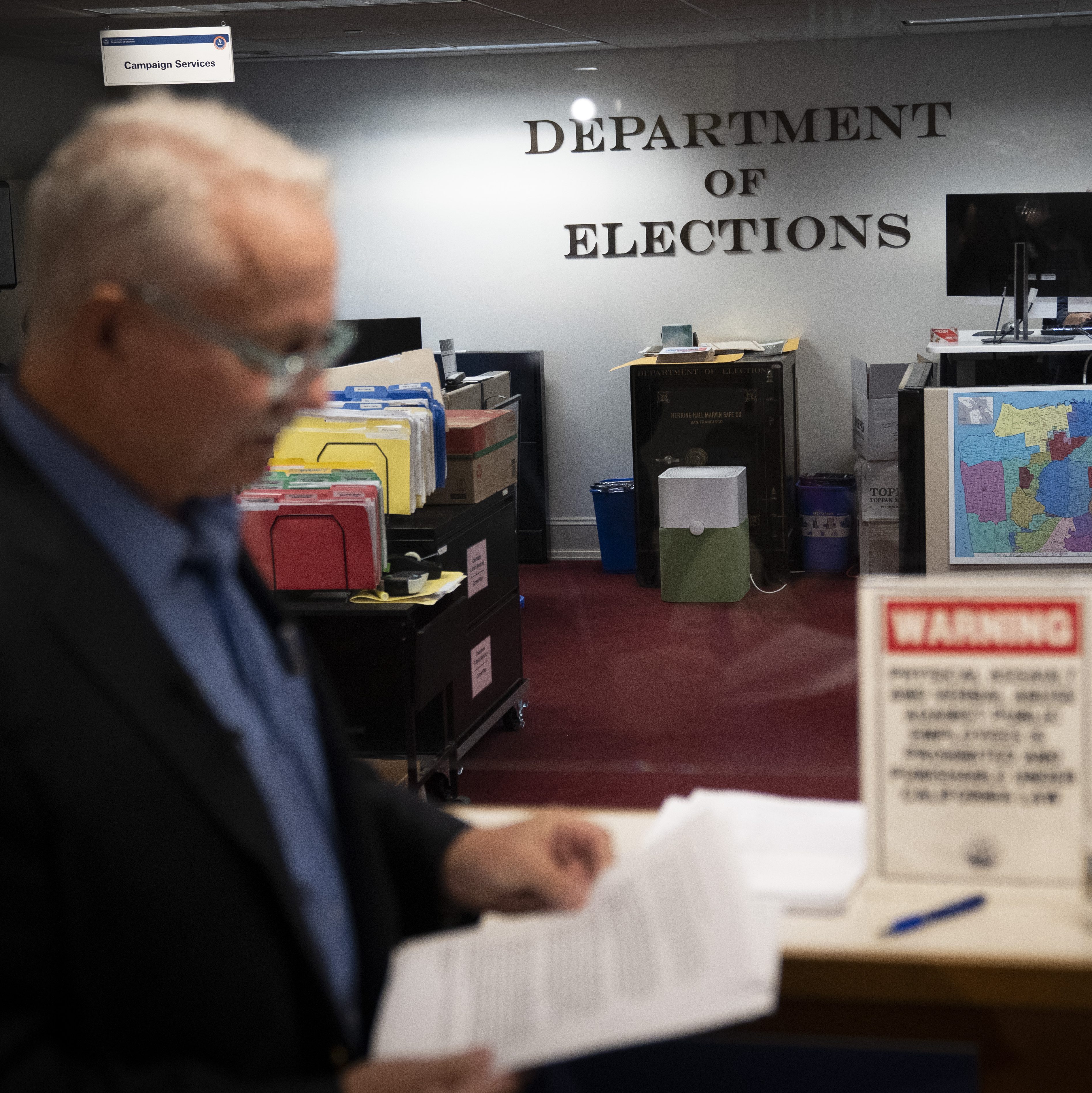 The image shows a blurred man in an office labeled &quot;Department of Elections,&quot; with organized file folders, a warning sign, and a colorful map on the wall.