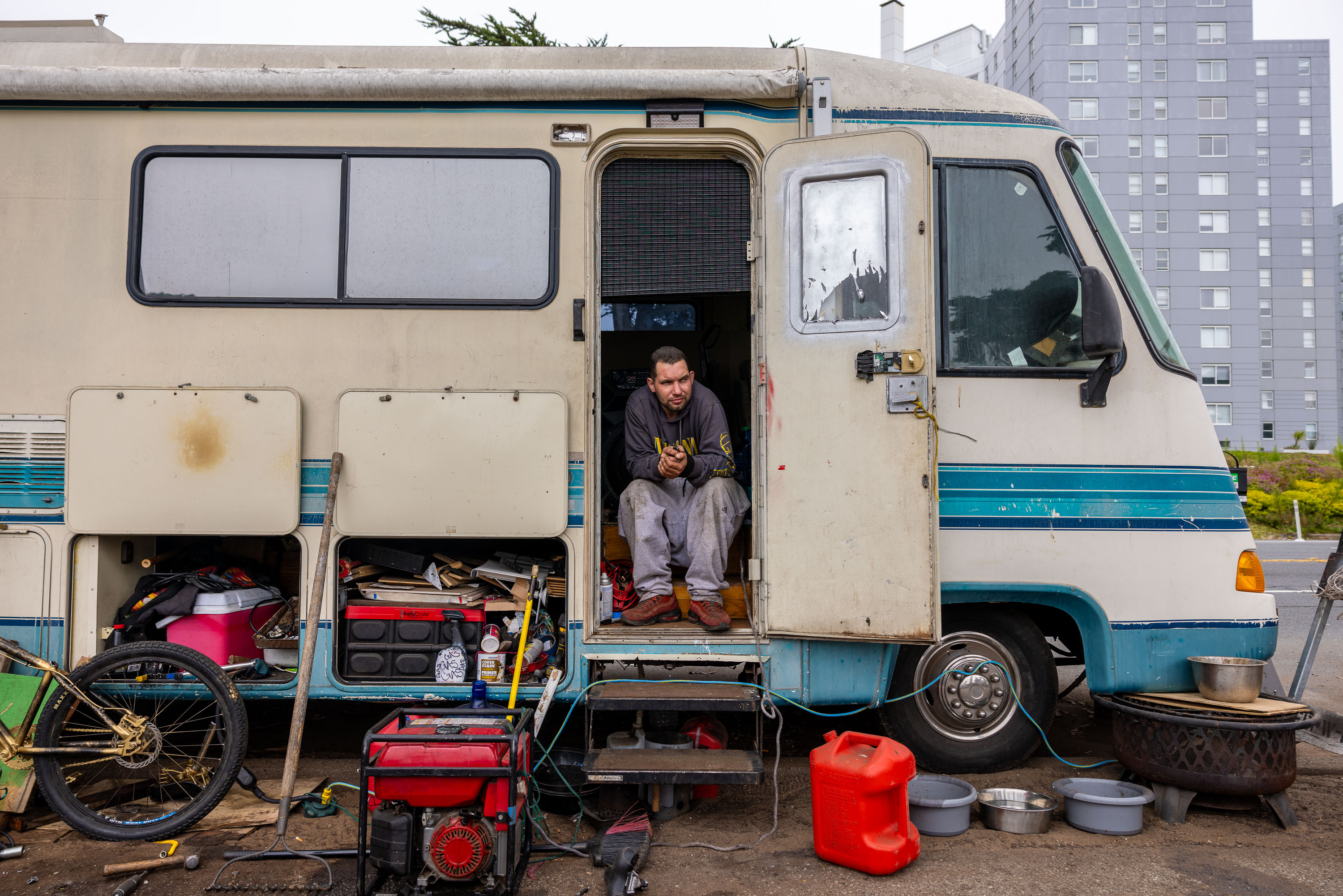 A man sits at the entrance of a cluttered RV parked on a street, surrounded by various items like a bicycle wheel, generator, and containers.