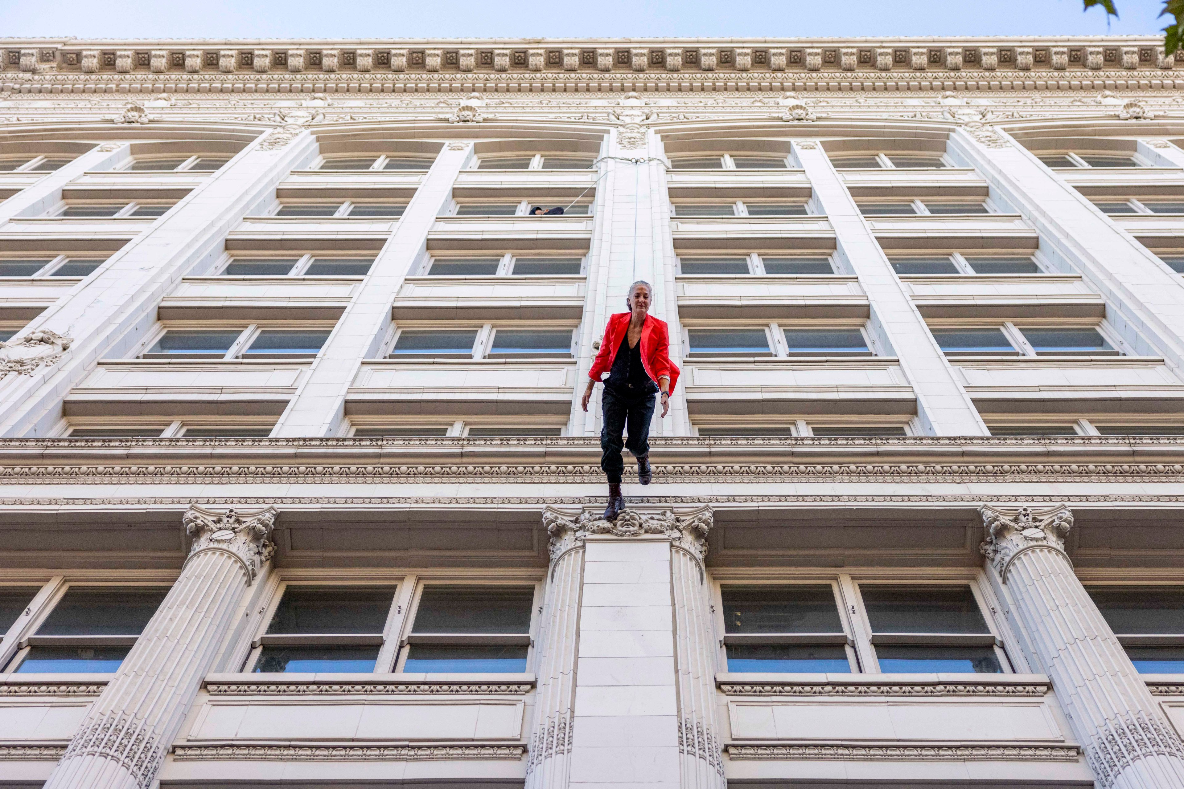 A person in a red jacket is dangling from the edge of a tall, ornate building with large windows and decorative columns, set against a clear blue sky.