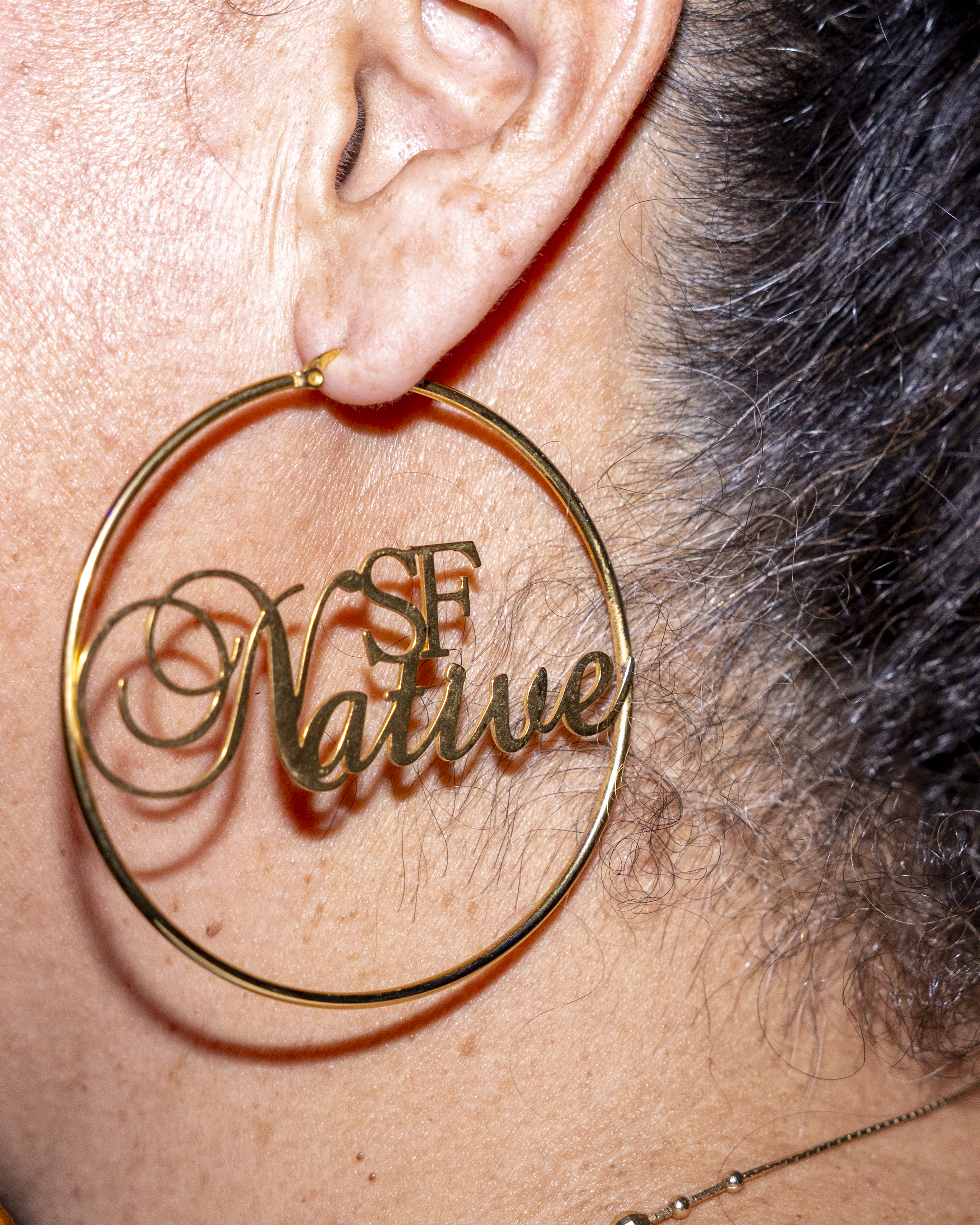 A close-up of an ear wearing a large gold hoop earring with the words &quot;SF Native&quot; scripted inside. The background shows hair and the earlobe.