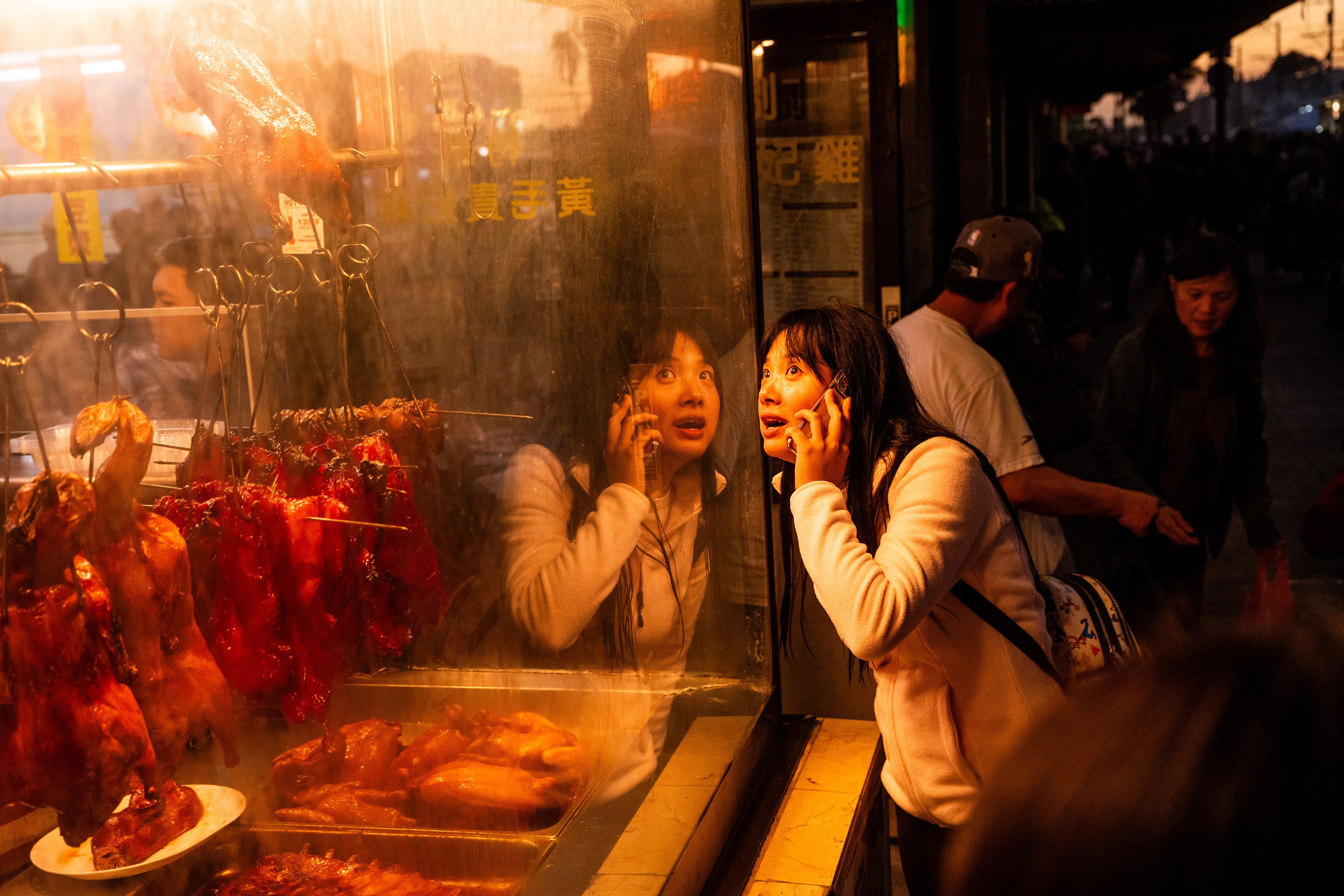 A woman talks on the phone while peering into a window displaying roasted meats. Her reflection is visible, and people walk by on the dimly lit street.