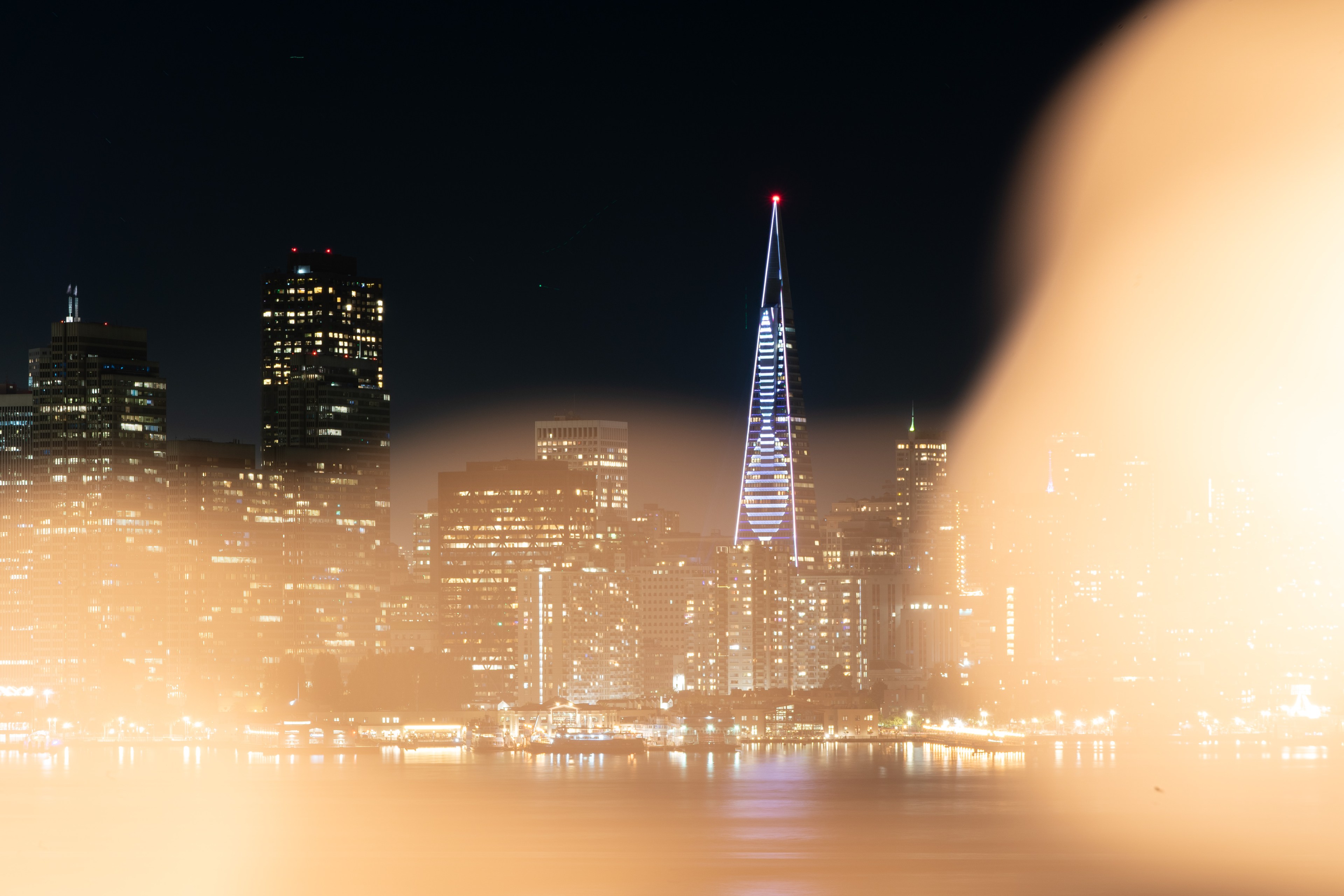 A city skyline at night features a prominent, illuminated triangular skyscraper. The foreground has a blurred, golden haze, likely from lighting or reflection.