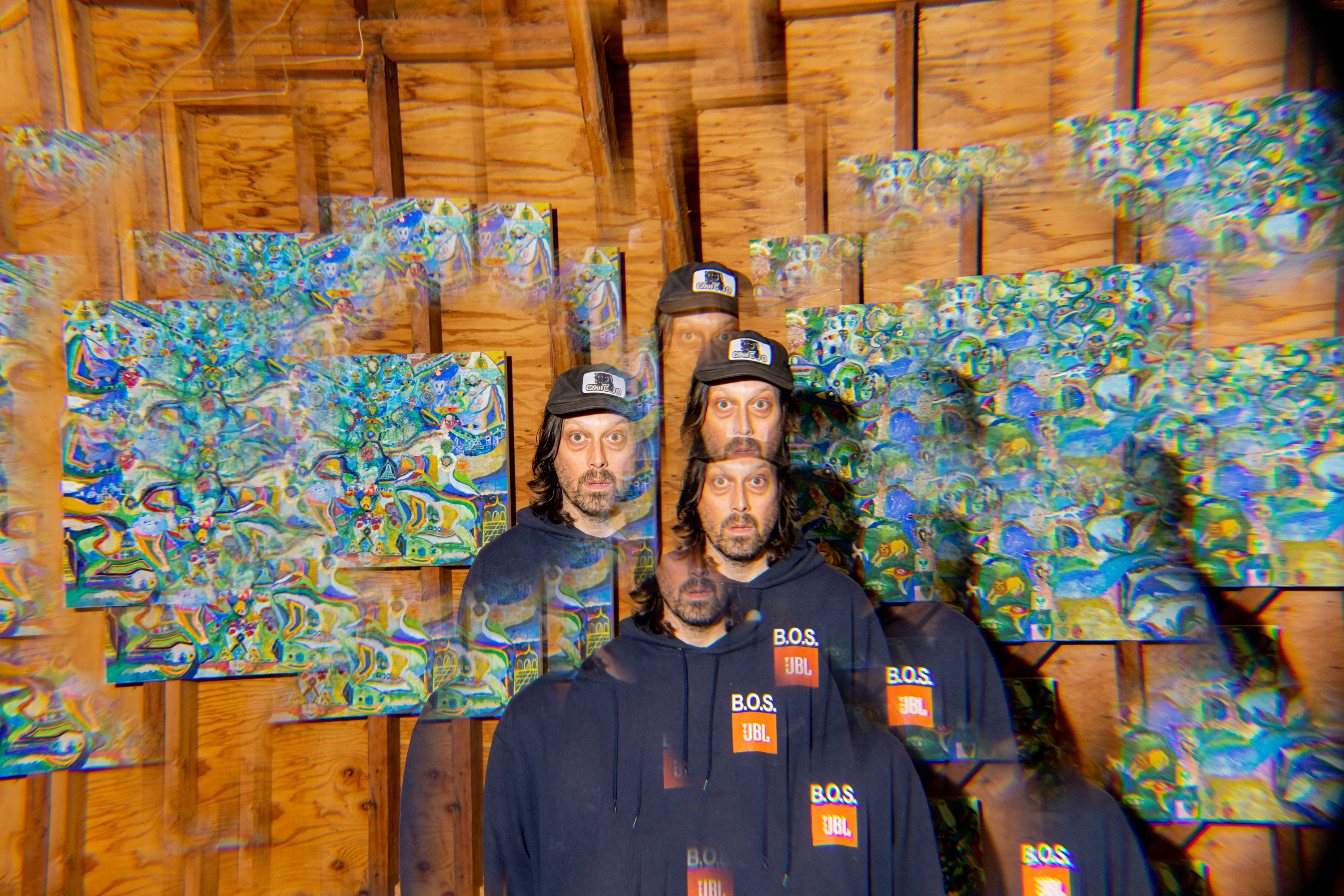 A man wearing a cap and hoodie is standing in front of colorful kaleidoscopic artwork. The image is distorted with multiple overlapping copies of him.