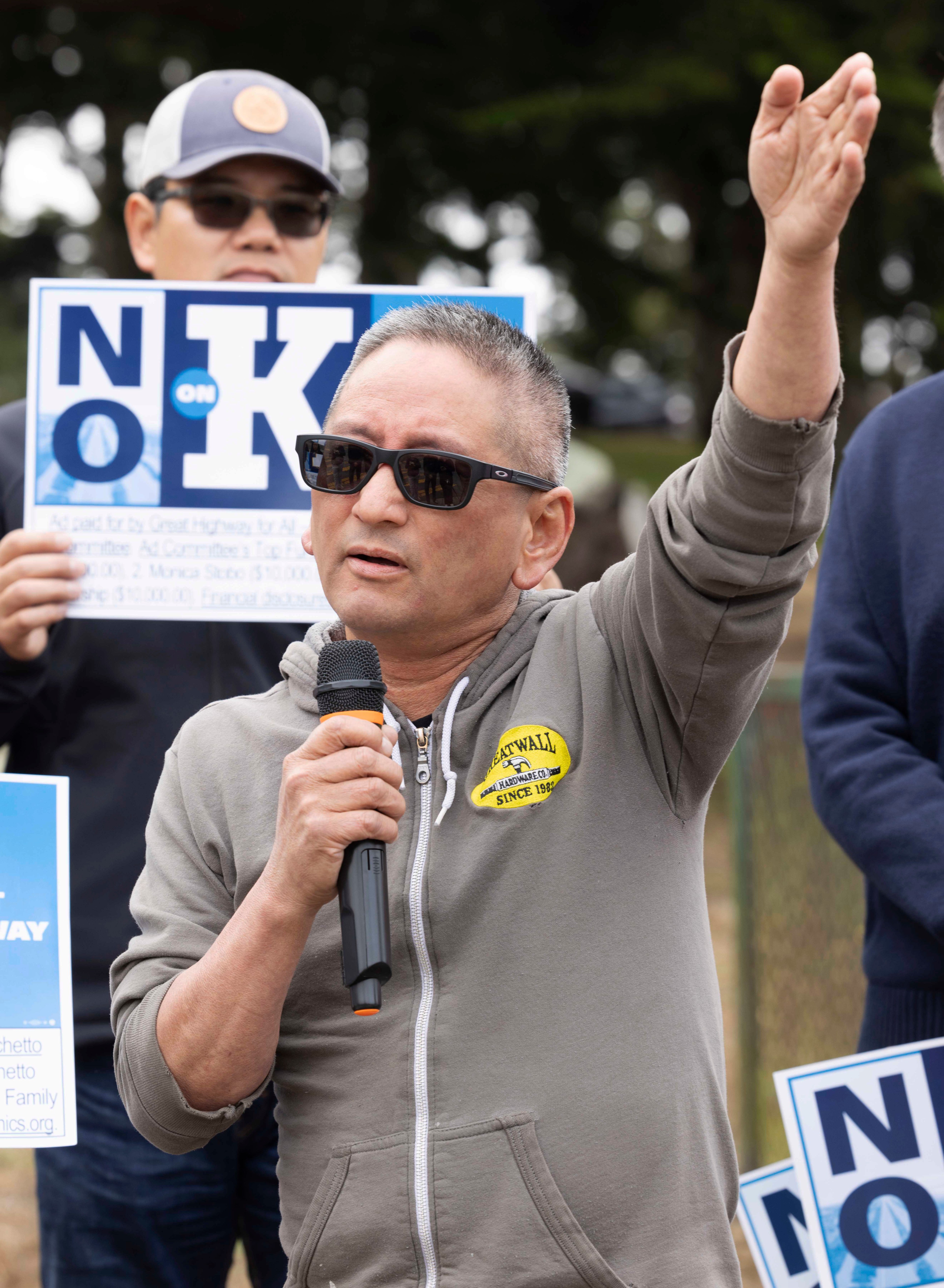 A man in sunglasses and a gray hoodie speaks into a microphone, gesturing with one hand. Behind him, another person holds a &quot;No on K&quot; sign.