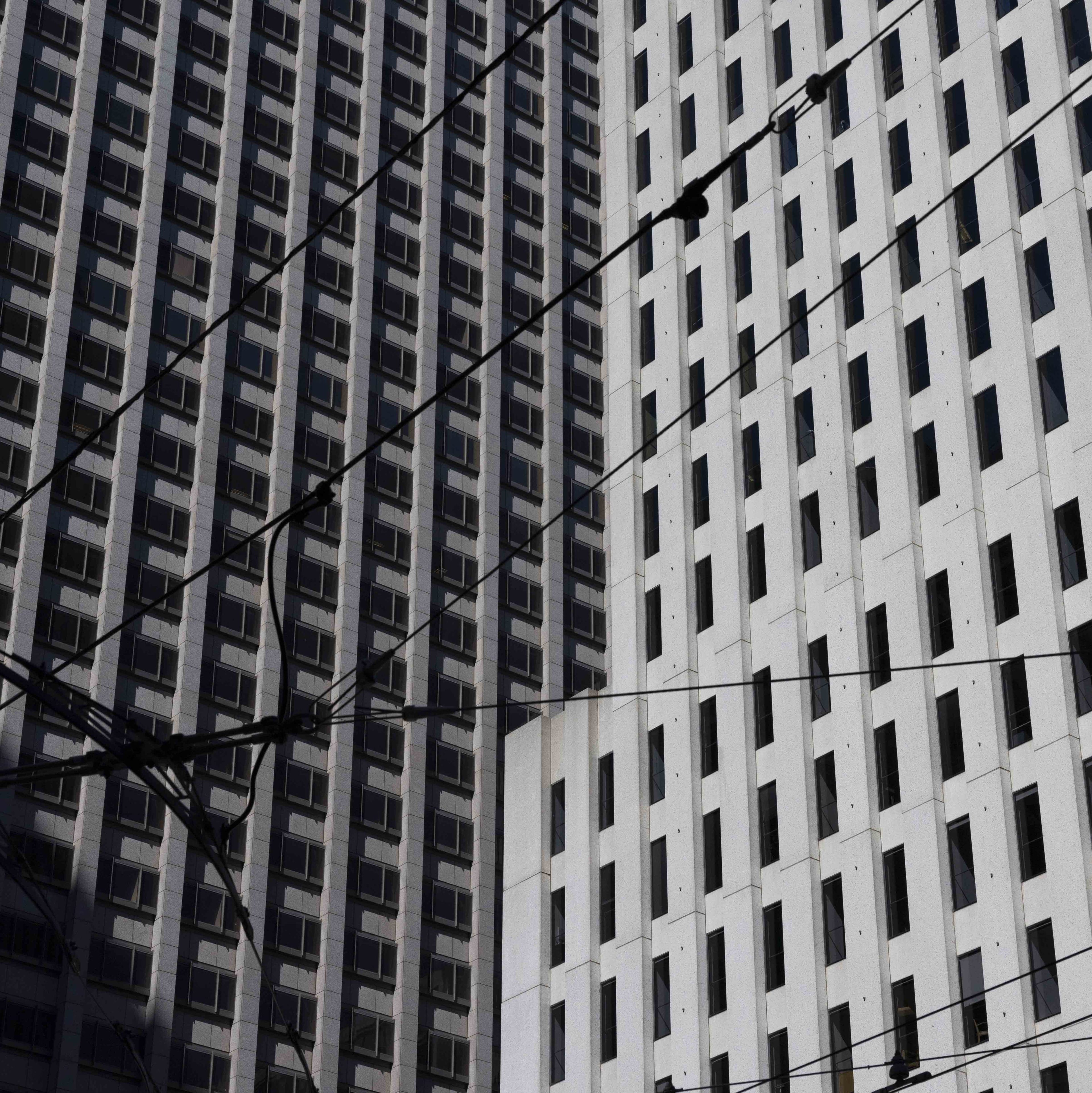 The image shows two tall buildings with many windows. The building on the left is darker, and the one on the right is lighter. Overhead power lines crisscross the scene.