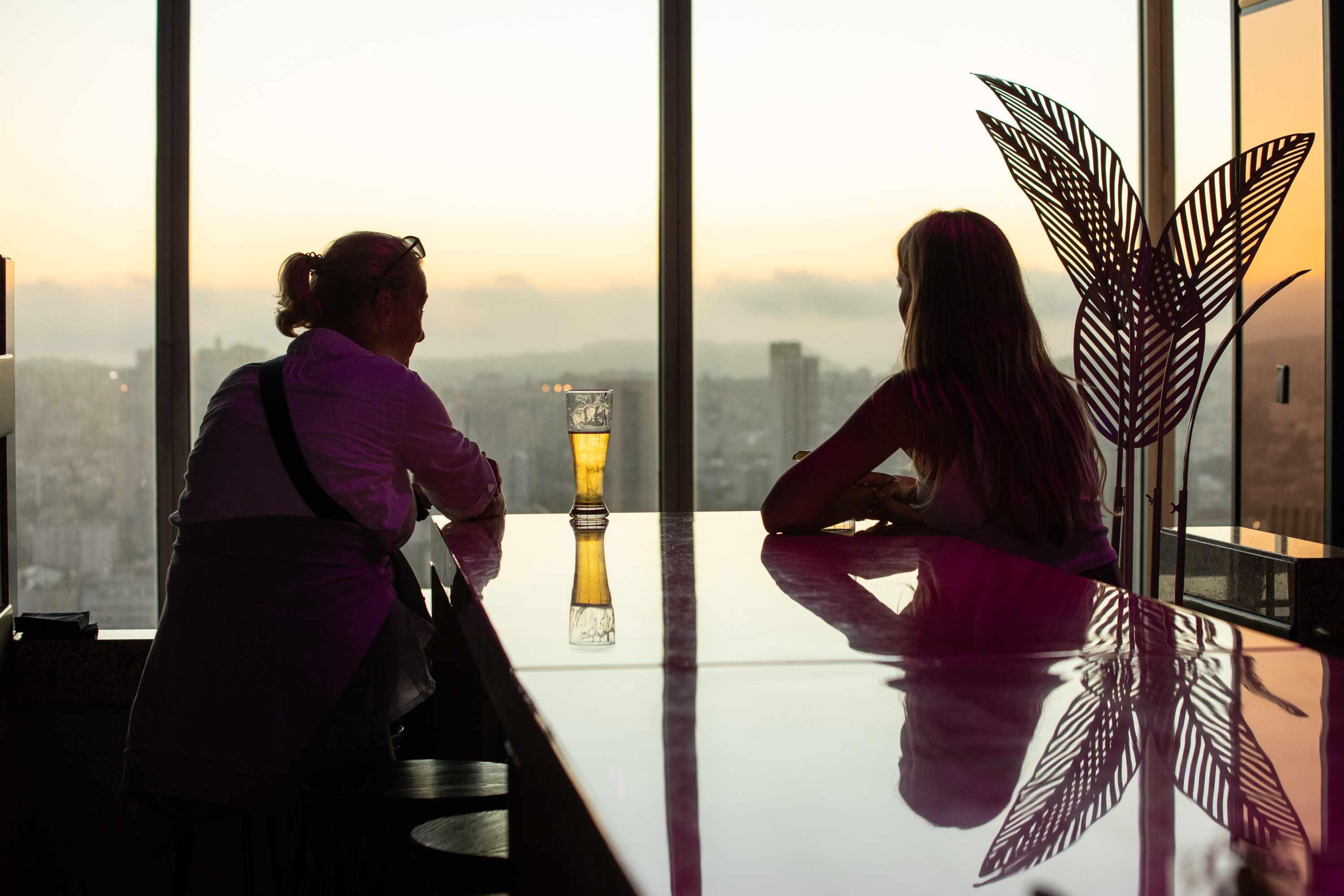 Two people sit at a bar with a clear window view of a city skyline at sunset. A drink rests on the glossy counter, reflecting the warm hues.