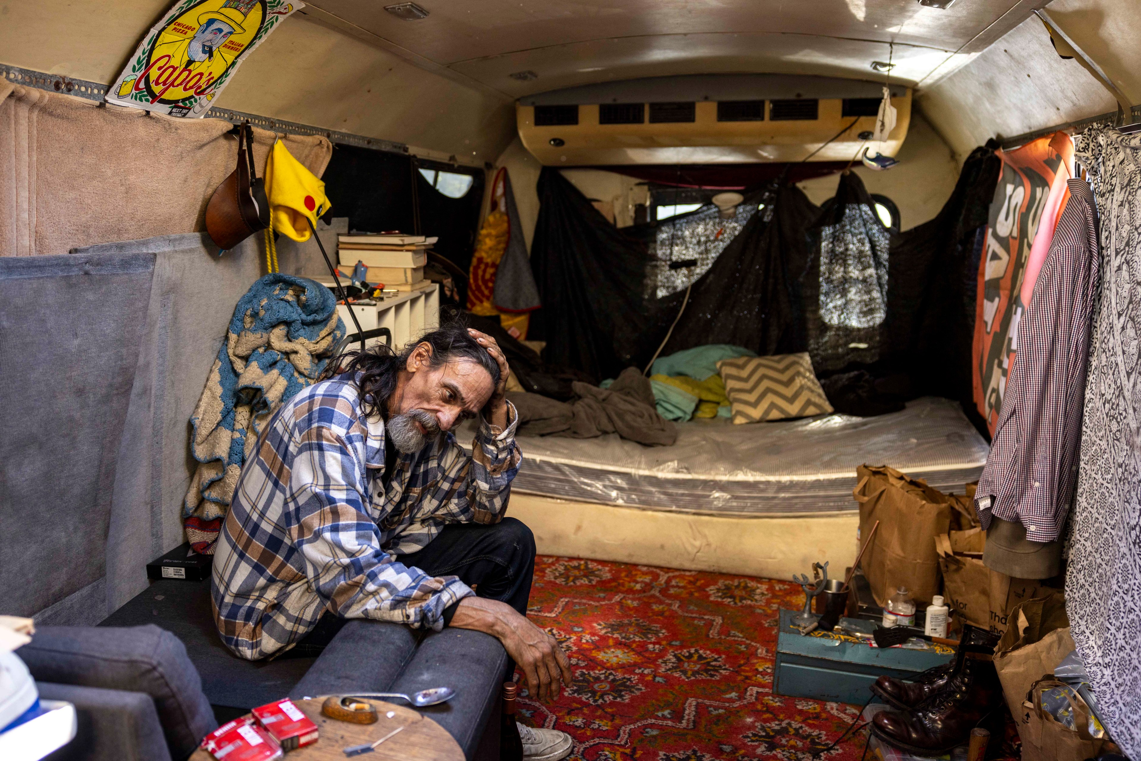 A man in a plaid shirt sits on a couch inside a cluttered trailer. The space features a sleeping area with pillows, various items scattered around, and a vibrant rug.