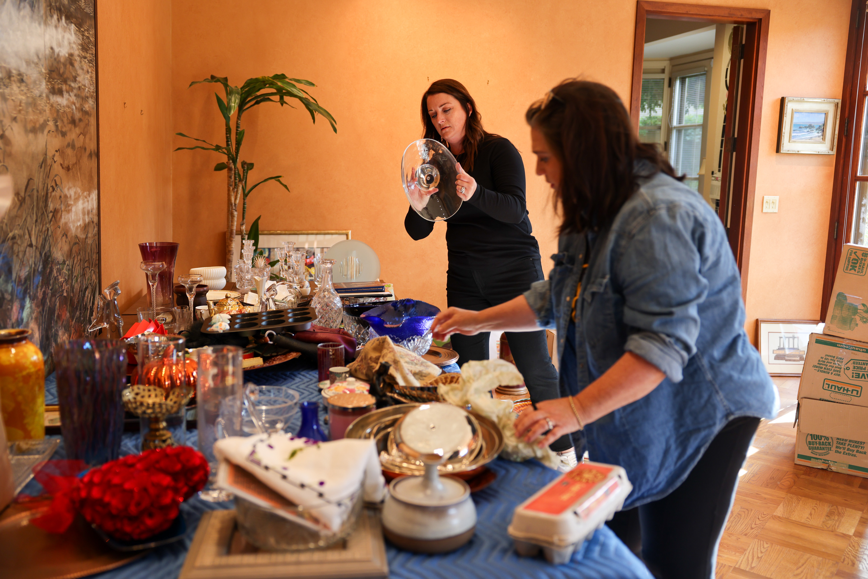 Two women sort through assorted glassware and decorative items on a table in a warmly lit room, surrounded by boxes and a potted plant.