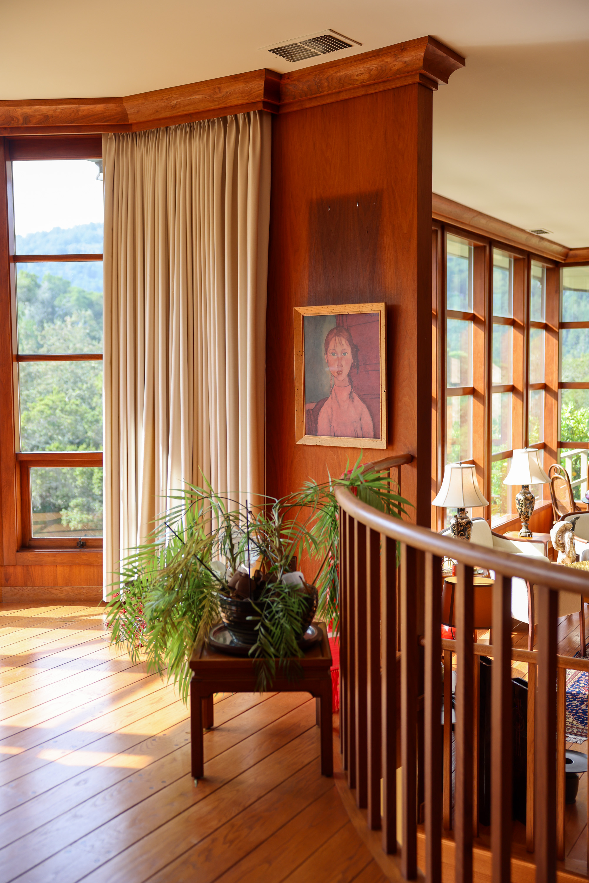 Sunlit room with wooden floors, large windows, a portrait on the wall, a potted plant on a table, white curtains, and two lamps on side tables.