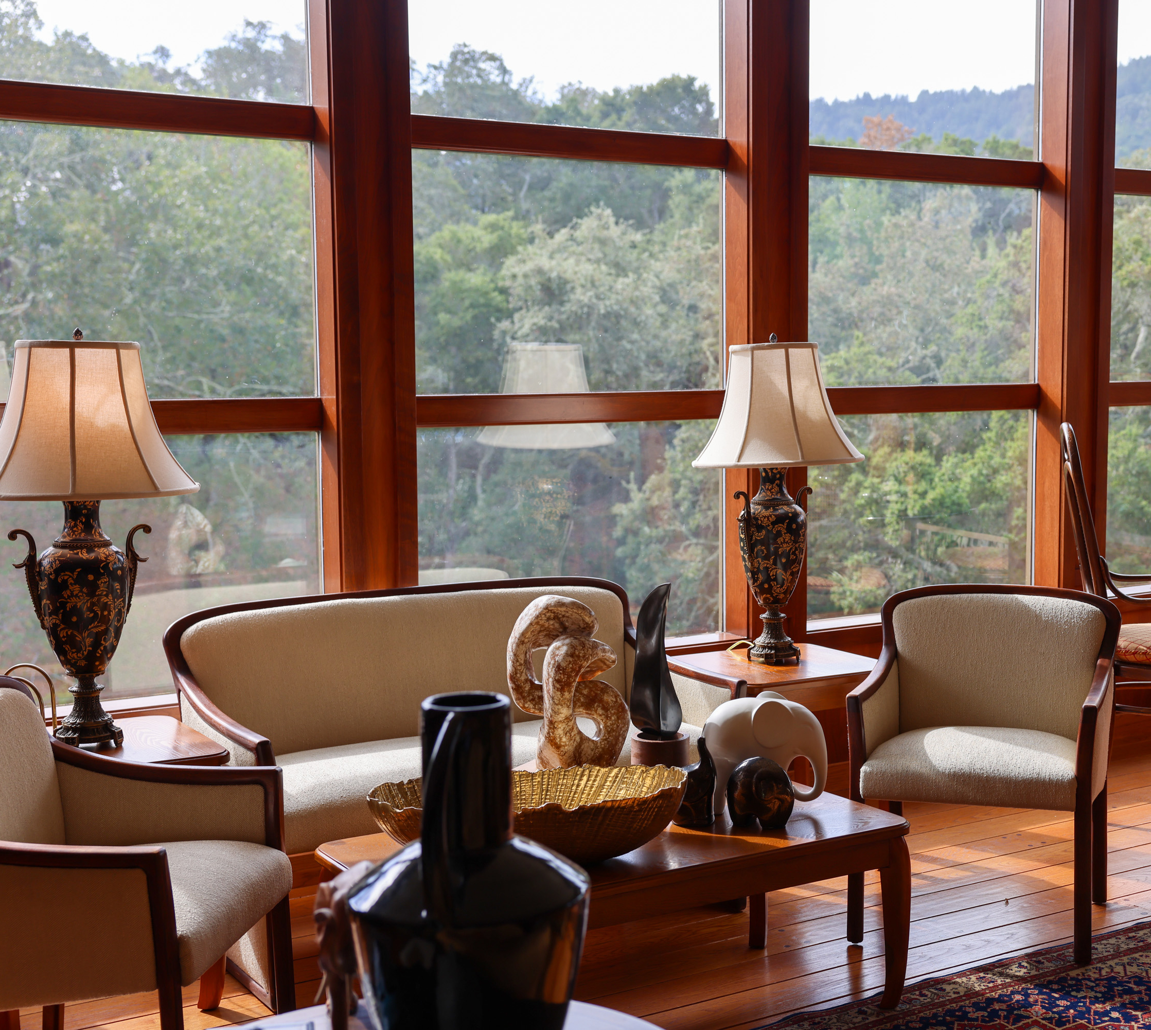 A cozy living room features beige chairs, a sofa, decorative lamps, and a wooden coffee table with sculptures. Large windows reveal a lush green view outside.