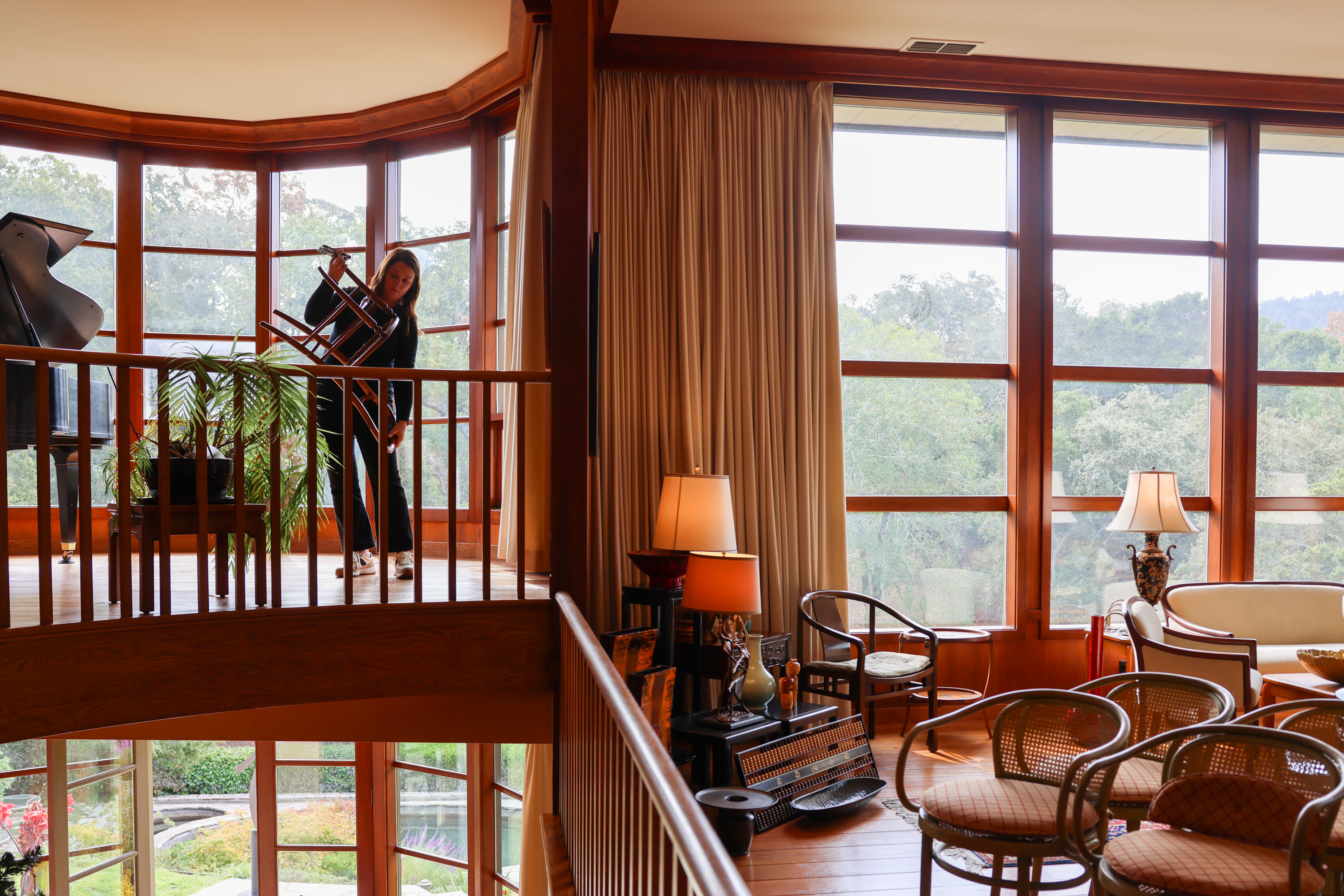 A room with large windows features a grand piano and plants on an upper level, while a cozy sitting area with lamps and chairs is seen below. A person adjusts a chair.