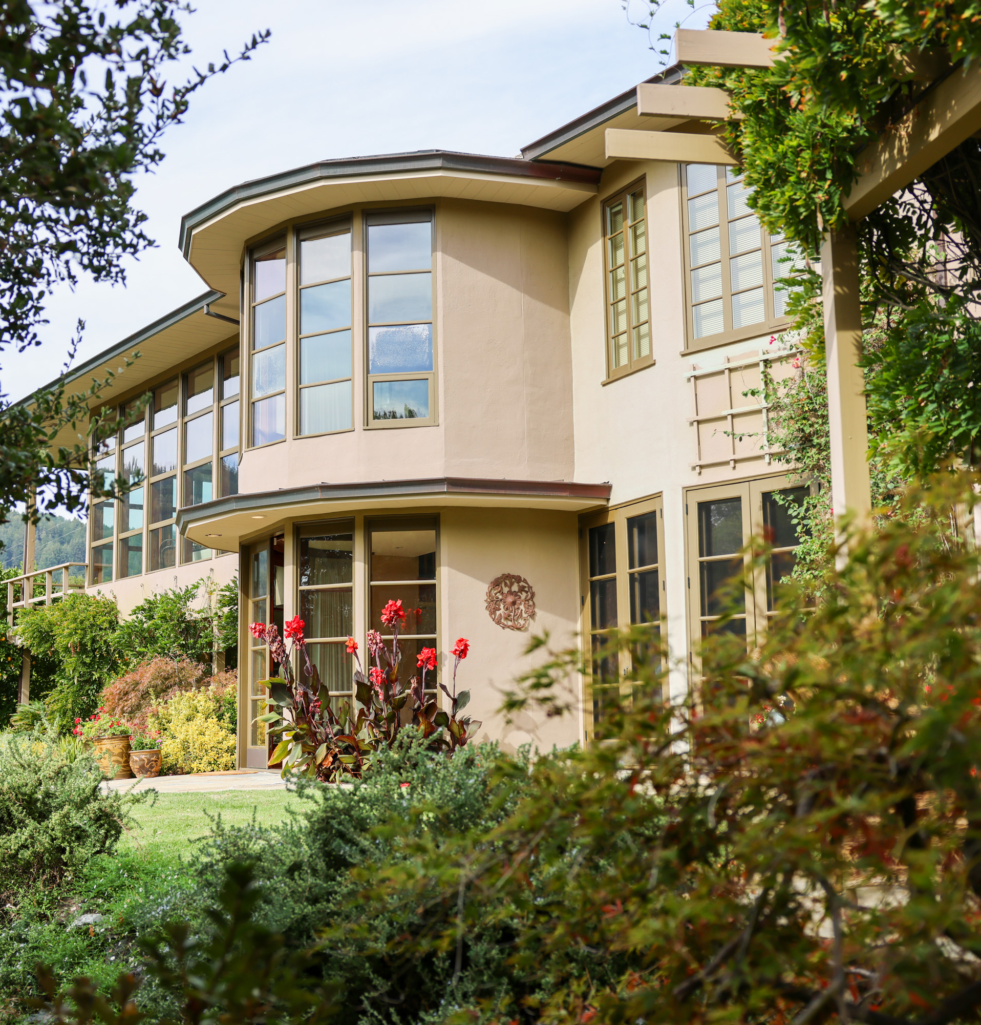 A modern house with large windows, cream walls, and a lush garden with red flowers and greenery. The structure features sharp, angular lines and a sunny atmosphere.