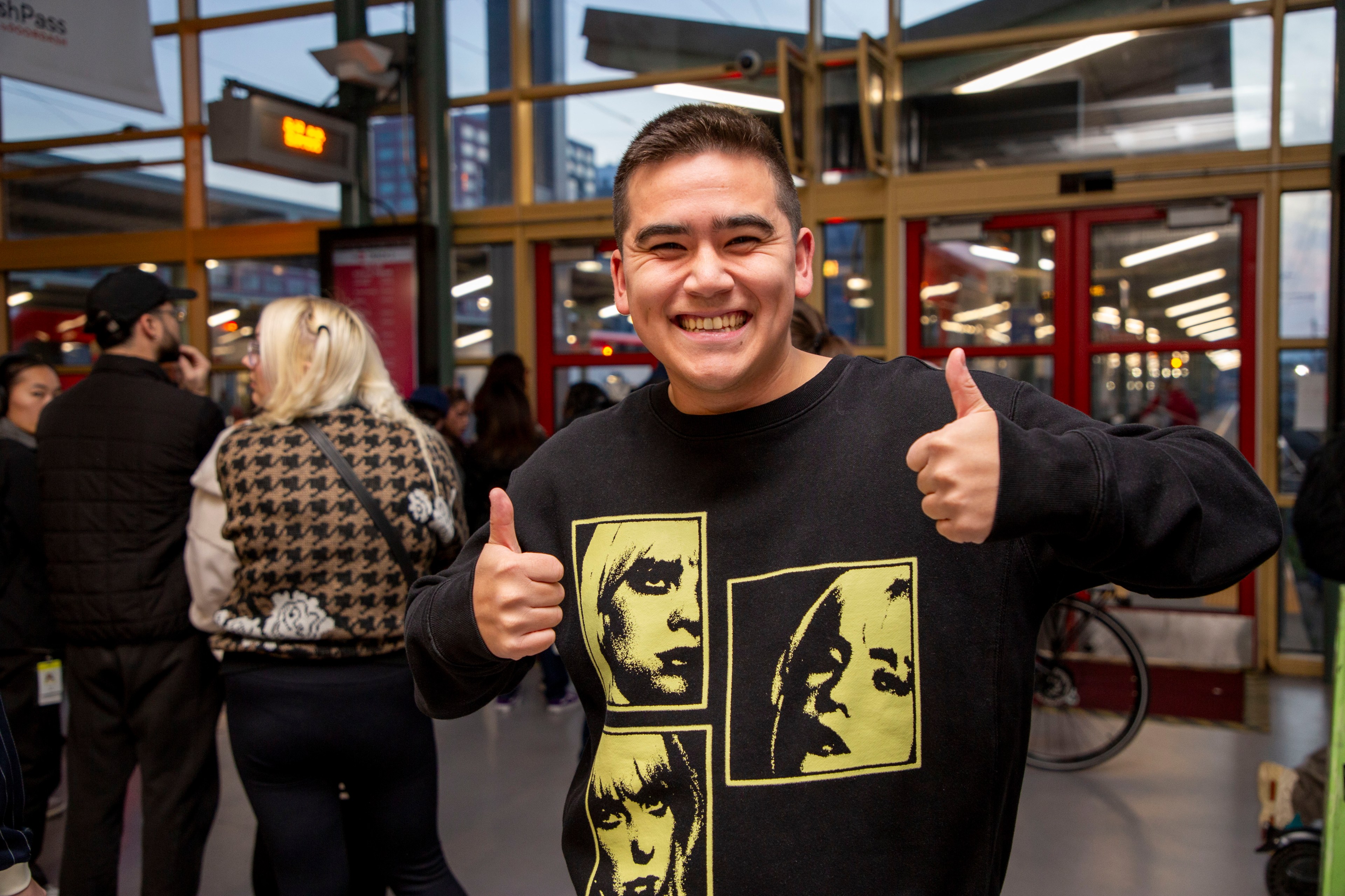 A smiling person wearing a black sweater with yellow graphic faces gives two thumbs up. They are standing indoors with people around and glass doors behind them.