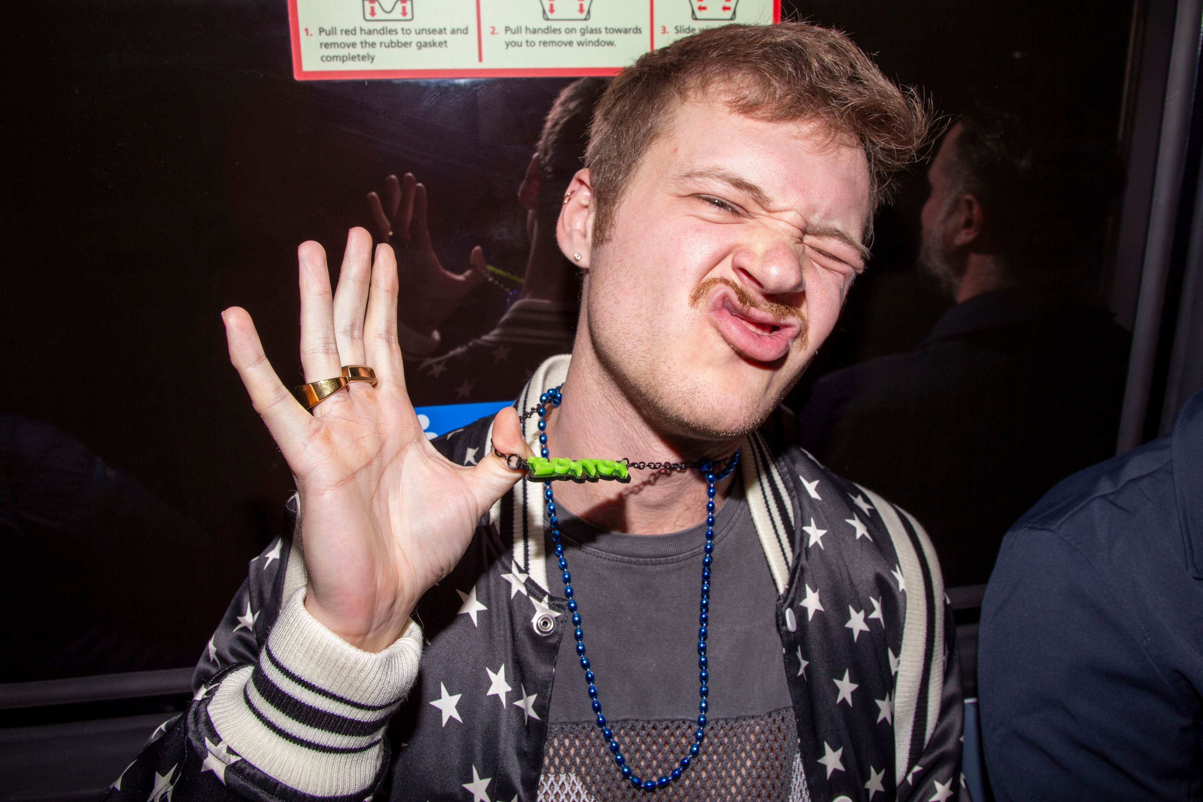 A person is making a playful face, holding a necklace with a green charm against a blue bead chain. They're wearing a star-patterned jacket.