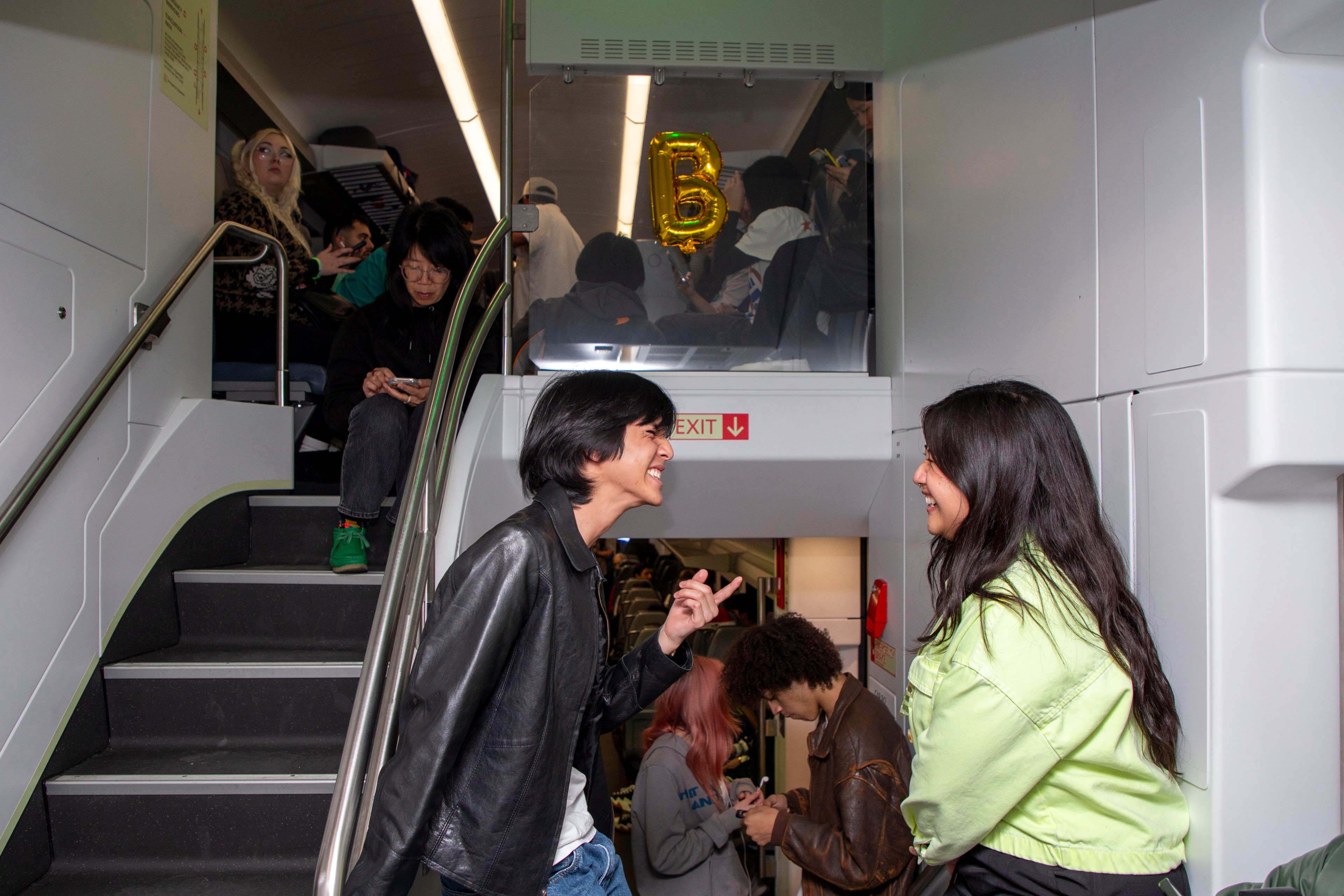 Several people are on a train, chatting and using phones. A staircase leads to an upper level where a gold “B” balloon is visible.