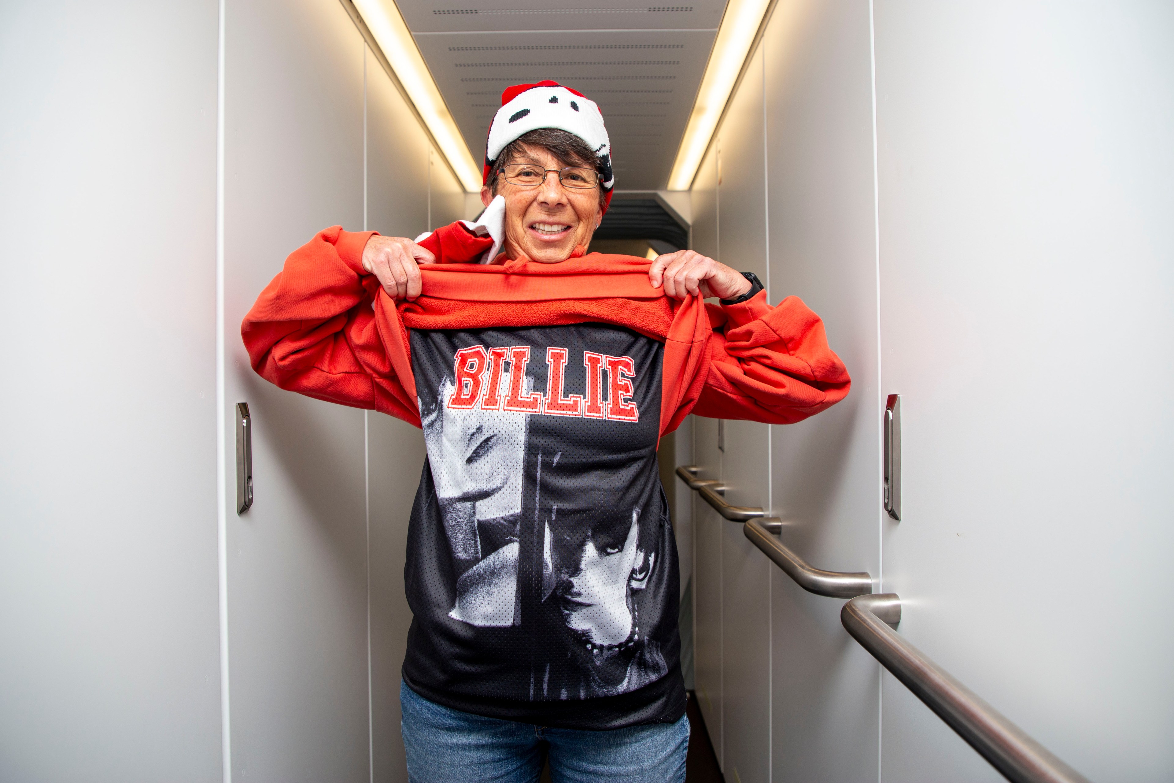 A person standing in a hallway lifts a red jacket to reveal a black shirt with &quot;BILLIE&quot; printed in red. They're wearing a Santa hat and glasses.