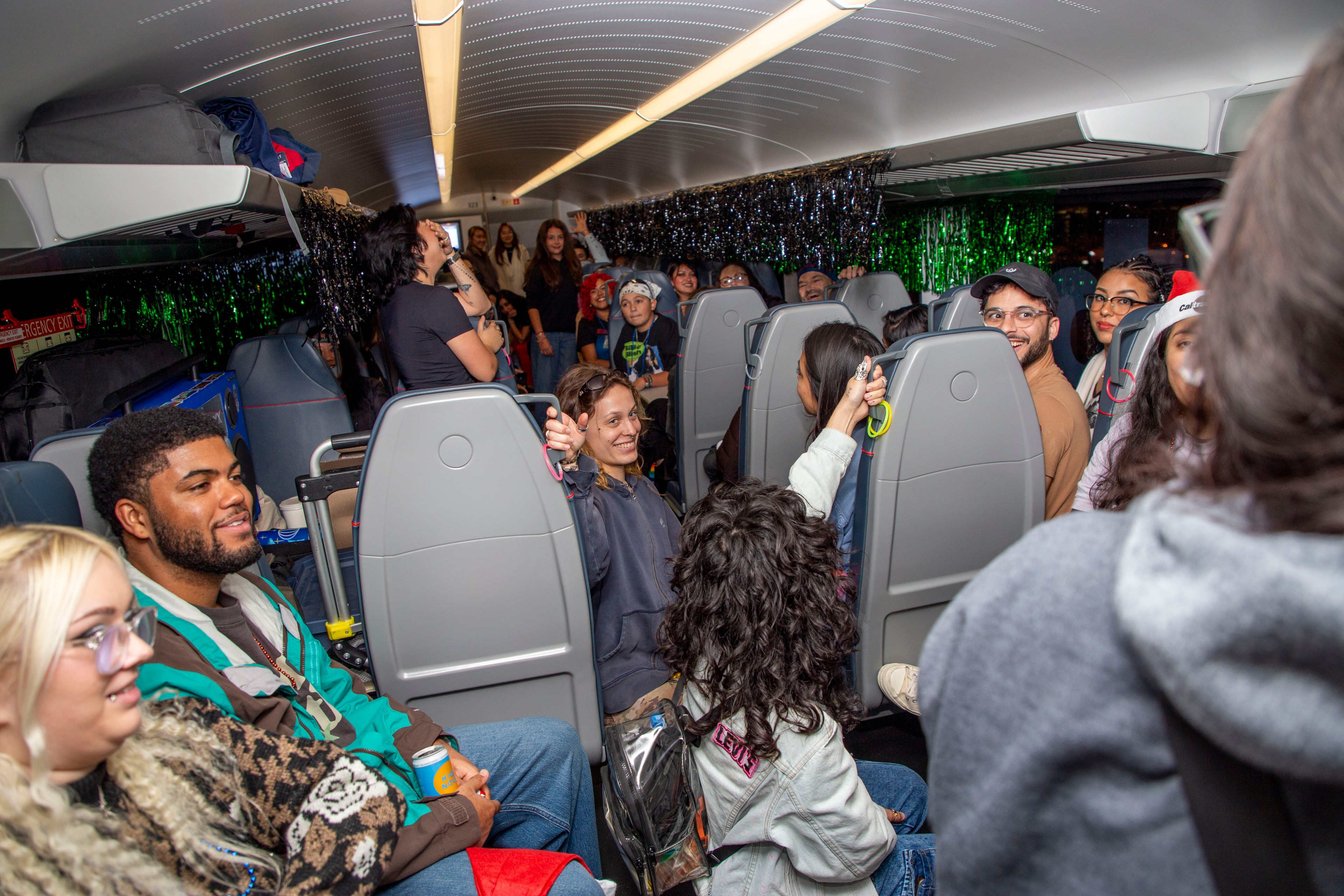 A group of people are sitting and standing inside a decorated train carriage, chatting and laughing. The shelves above hold bags, and the space feels lively and festive.