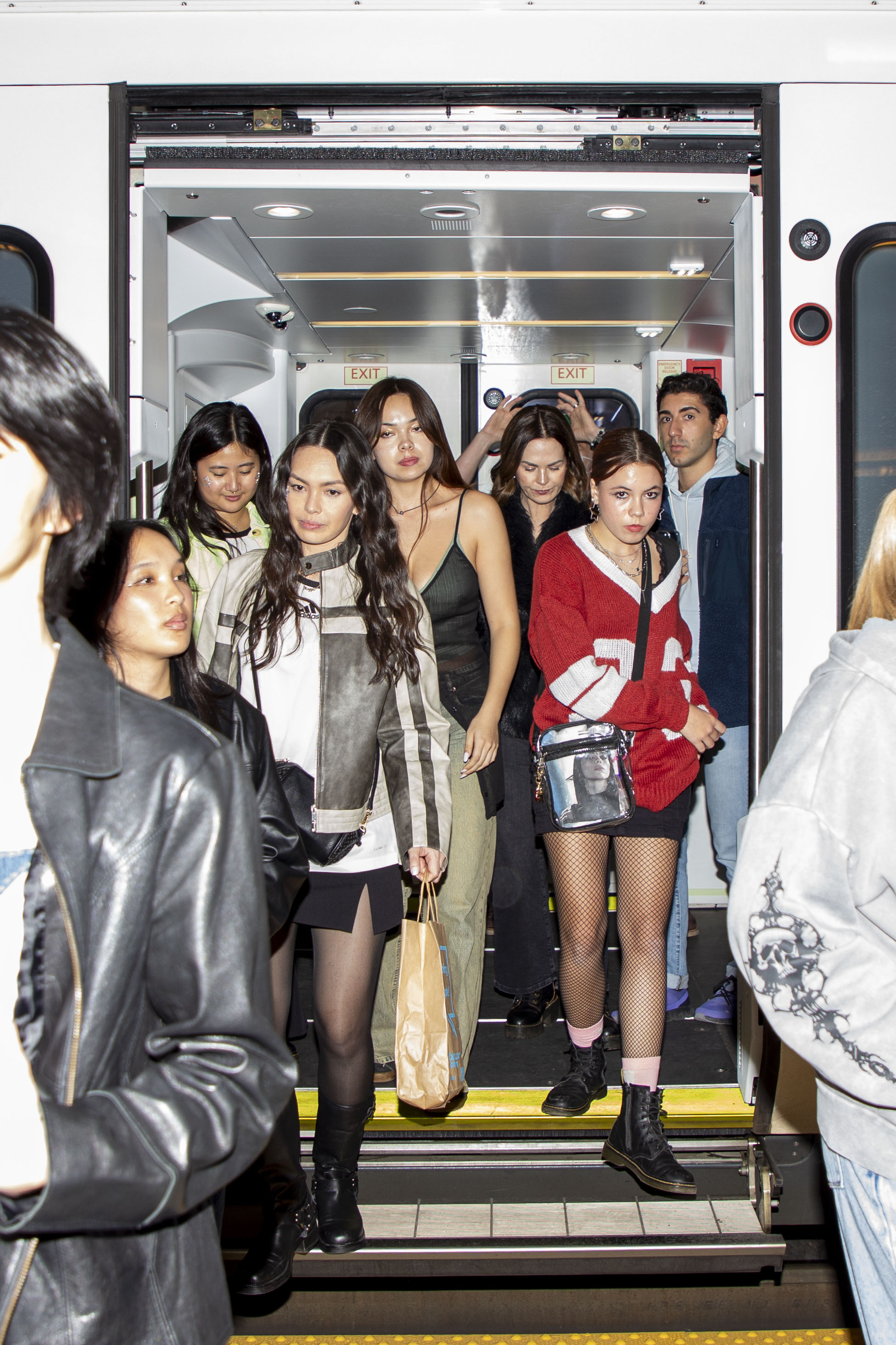 A group of young adults, dressed in trendy outfits, are exiting a train. They appear to be heading out for an event, carrying bags and looking lively.