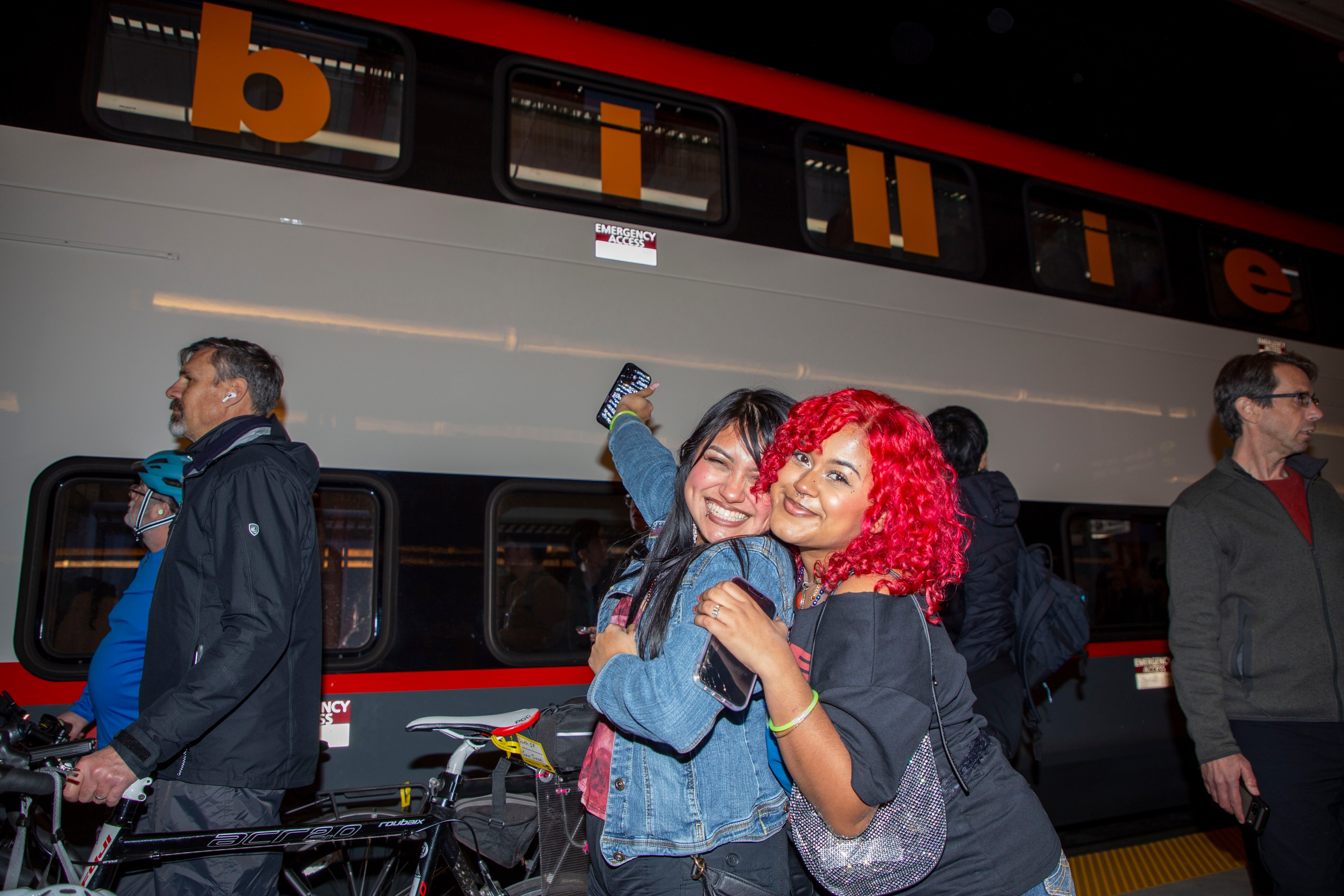 Two women smiling, one with red hair and one in a denim jacket, pose happily in front of a train. Other commuters and bicycles are nearby.