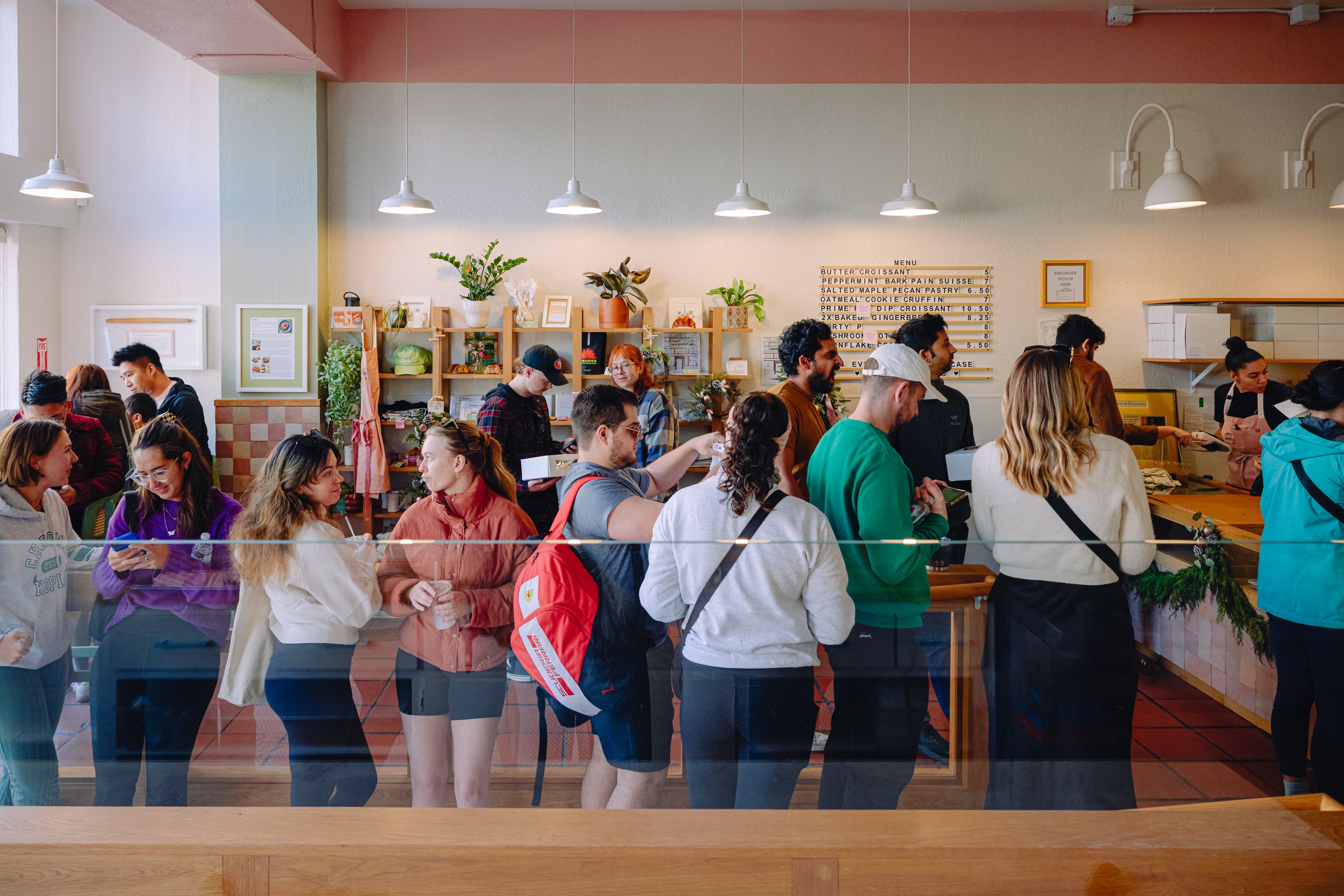 The image shows people waiting in lines both inside and outside a bakery. Inside, some are chatting and smiling, while outside, others stand patiently.