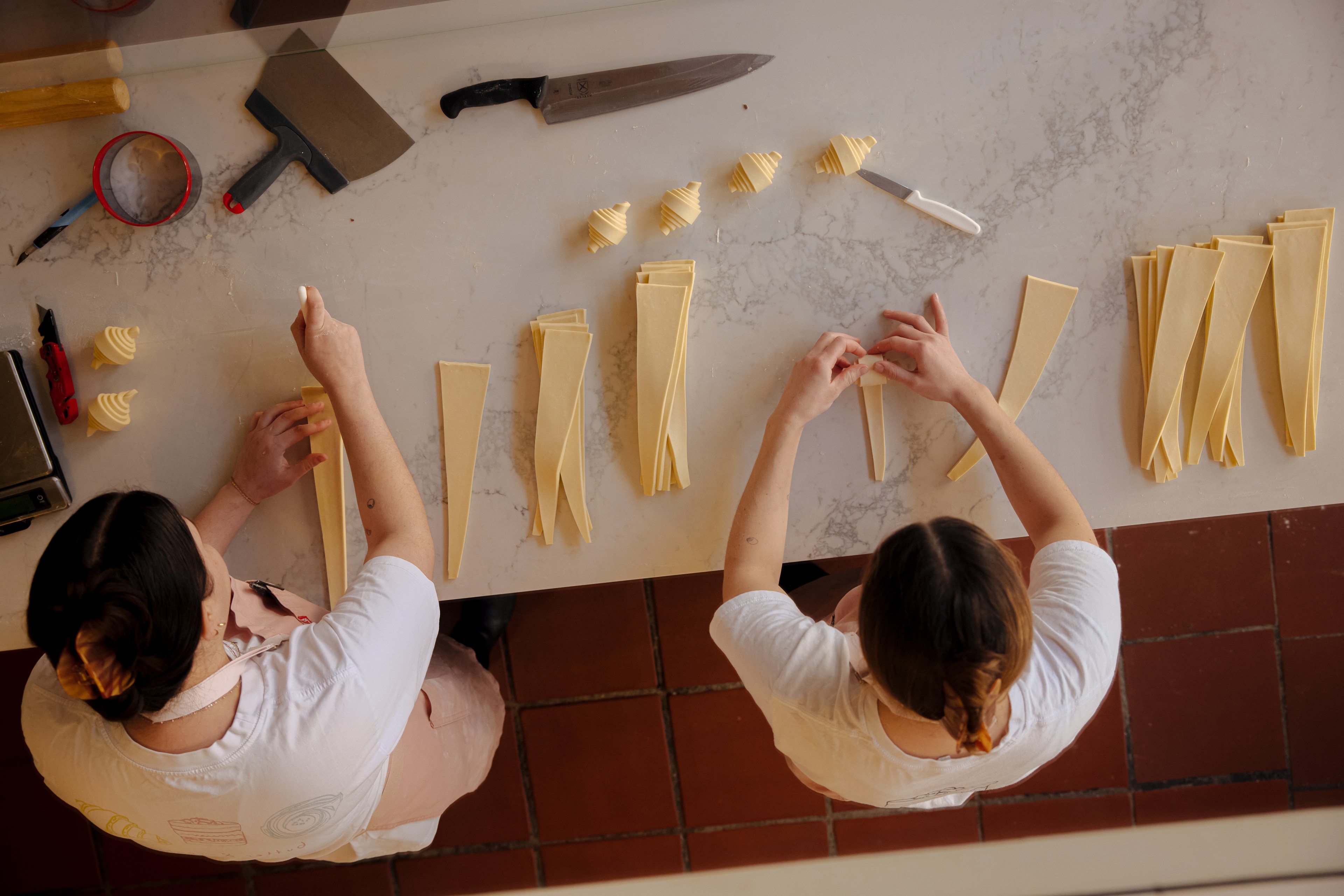 Two people are making pastries by folding dough strips on a marble countertop, surrounded by tools like a rolling pin, knives, and a scale.