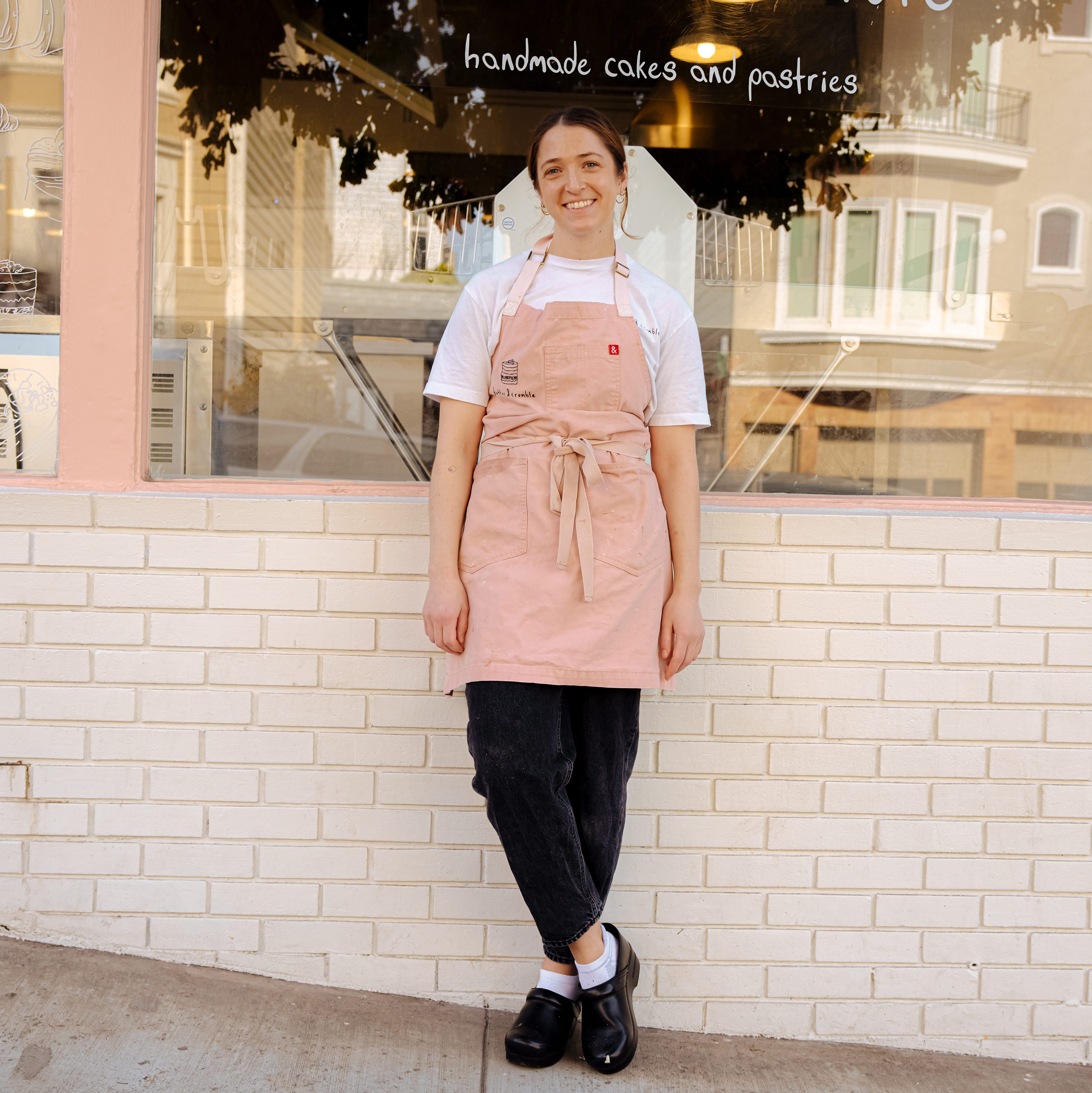 A person stands smiling in front of a bakery. They're wearing a pink apron over a white shirt and dark pants. The window displays &quot;handmade cakes and pastries.&quot;