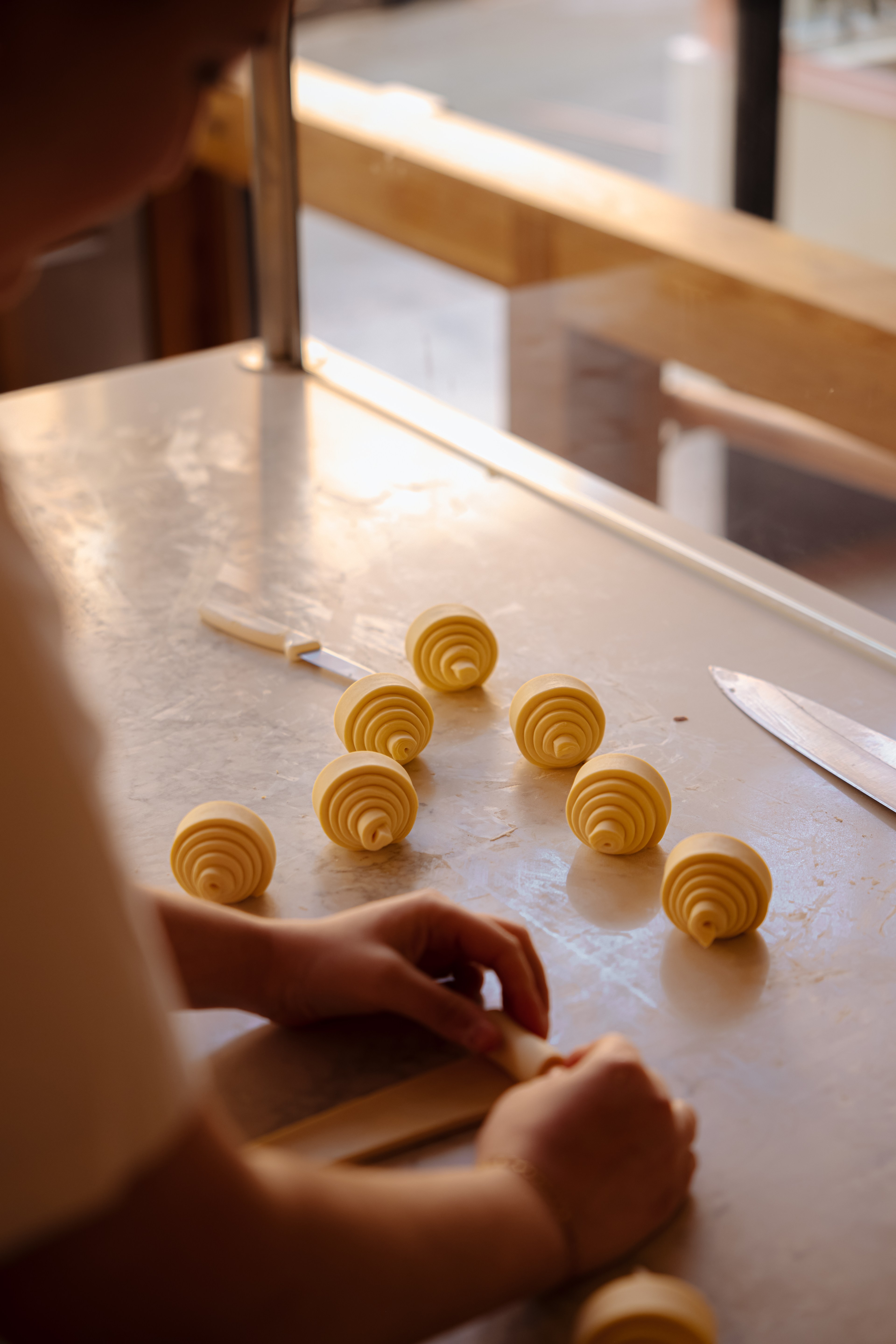 A person is shaping pastry dough on a counter. Seven spiraled dough pieces are neatly arranged next to a knife in a sunlit kitchen area.