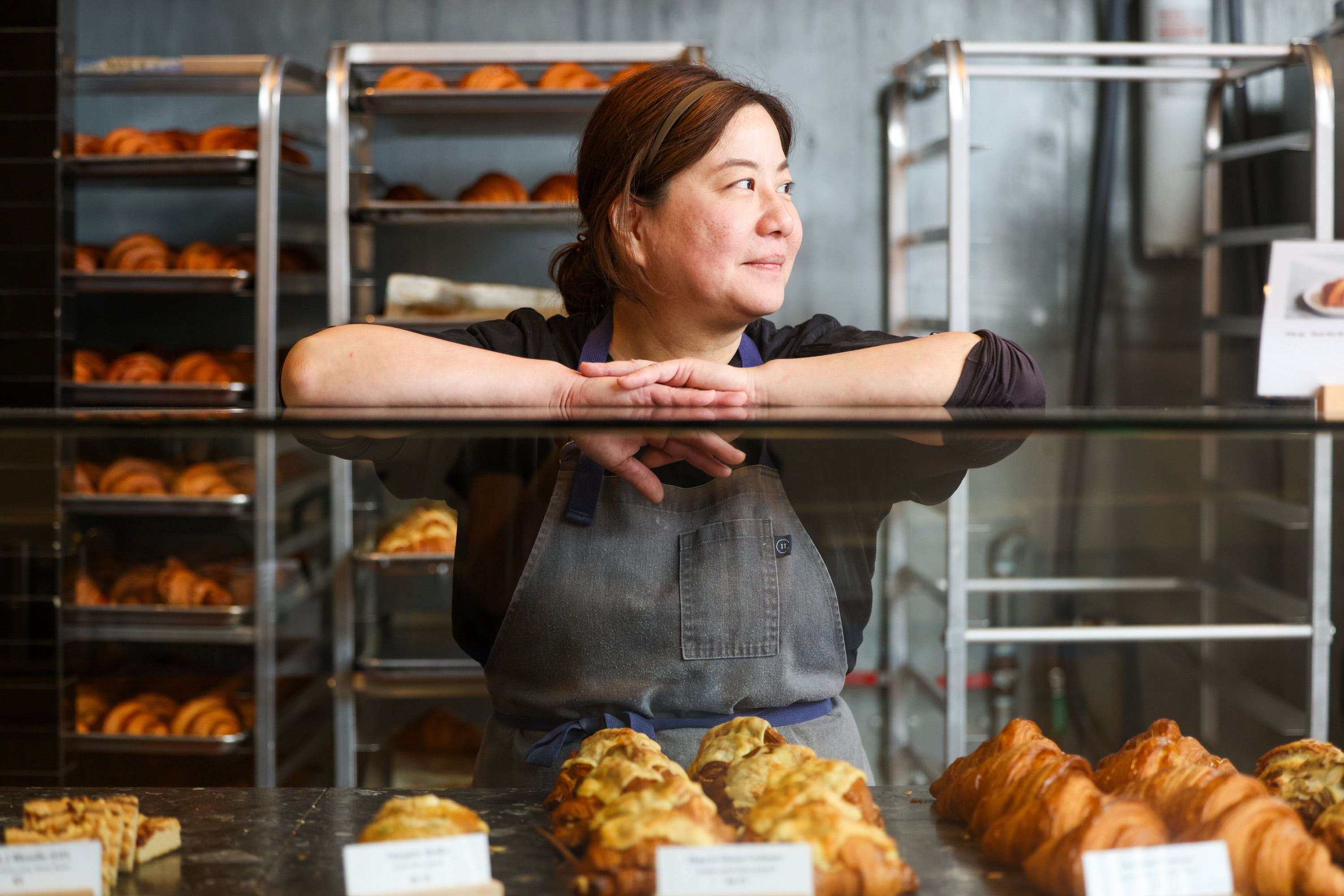 A woman in an apron stands behind a bakery display case, looking sideways. The counter features croissants, and bakery racks with pastries are in the background.