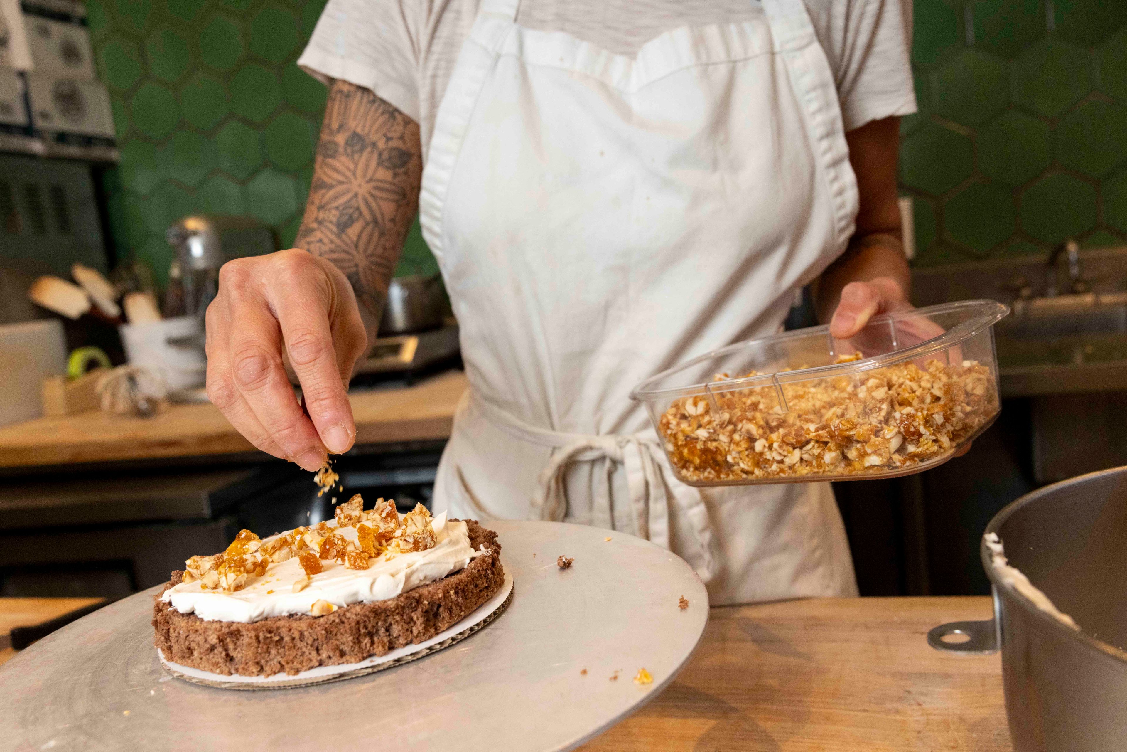 A person wearing a white apron is decorating a cake with caramelized nuts. They have a tattooed arm and are placing toppings on cream-covered brown cake.