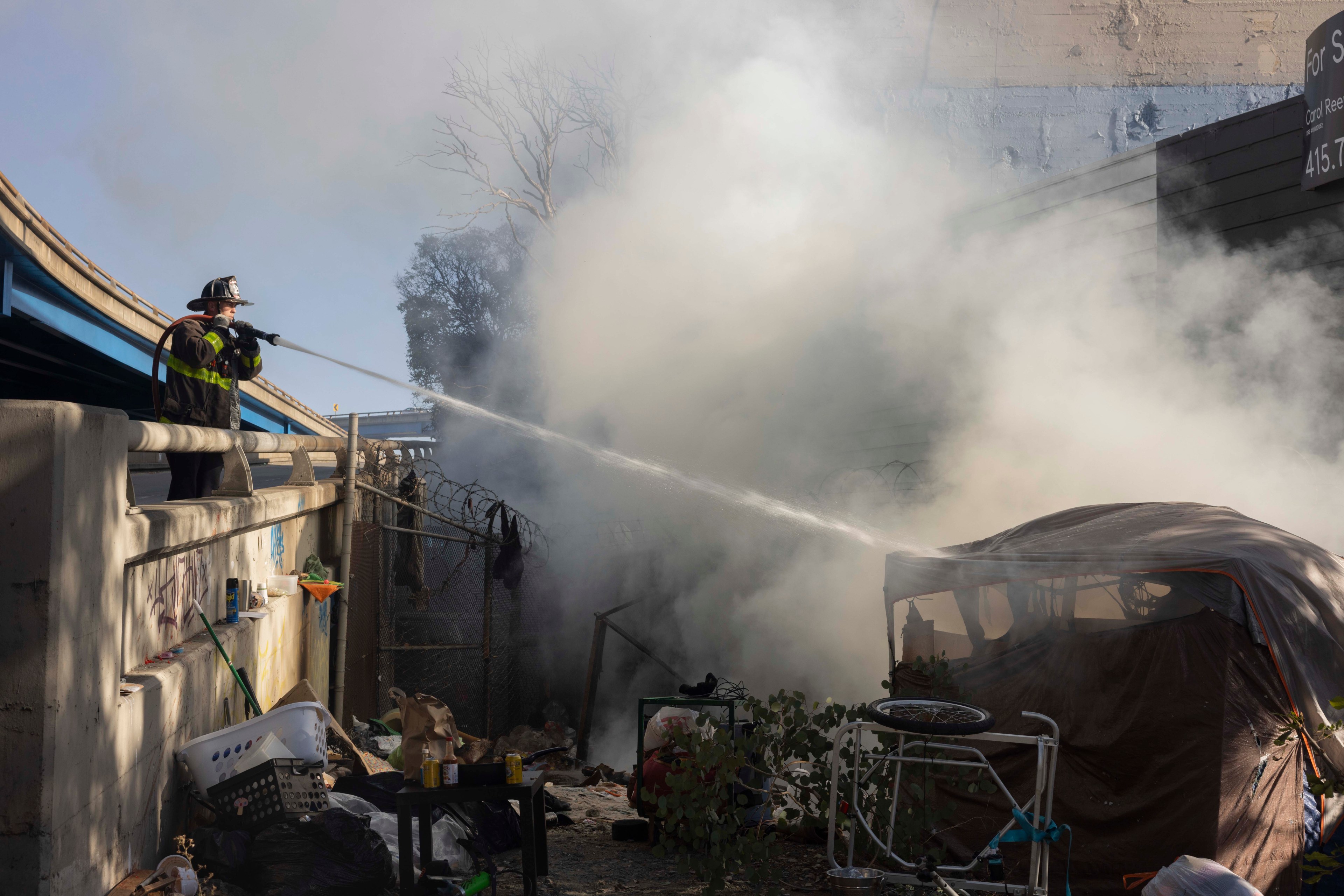 A firefighter sprays water onto a smoky area near a makeshift tent encampment under an overpass, surrounded by scattered items and debris.