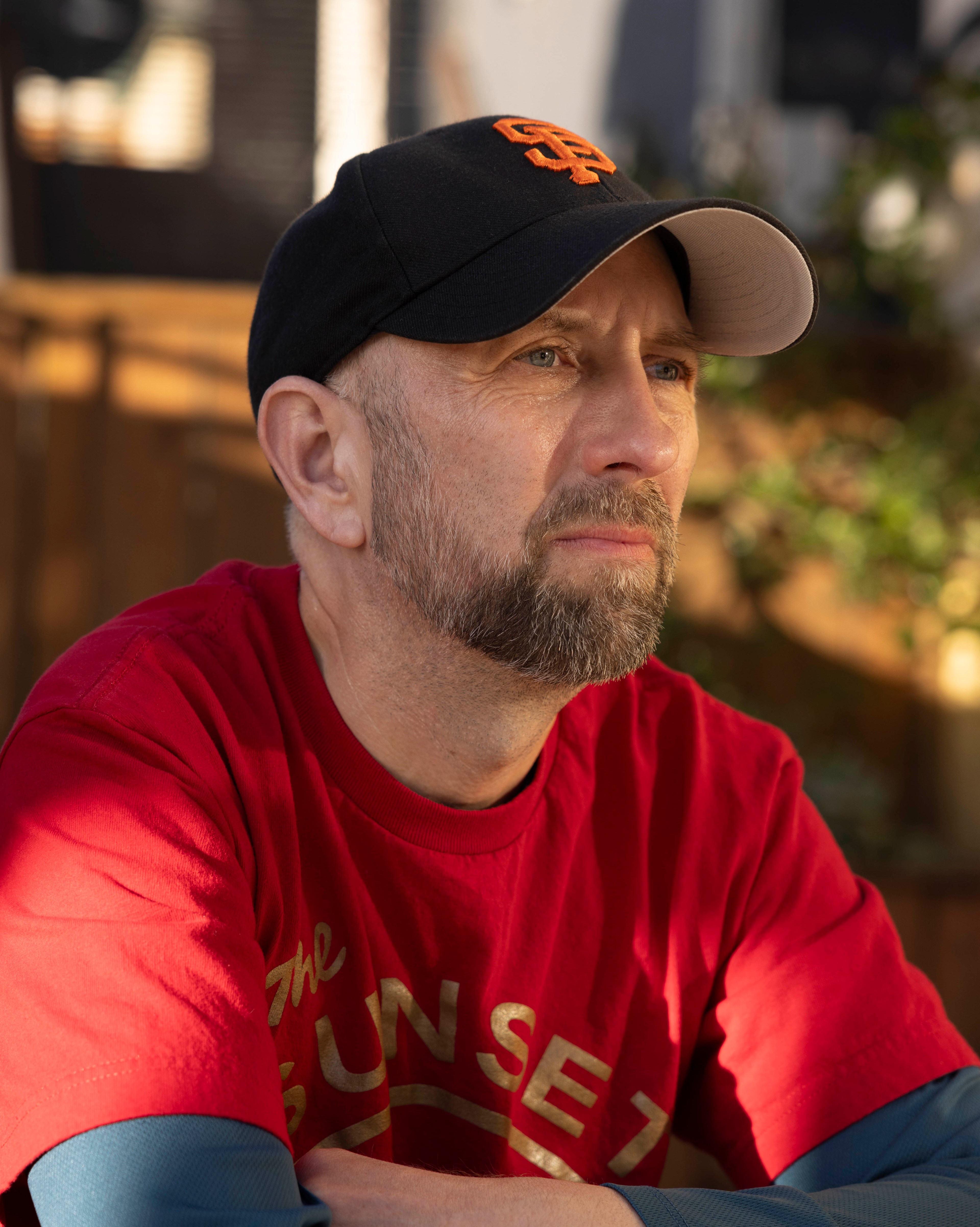 A bearded man in a black baseball cap with orange lettering gazes thoughtfully into the distance, wearing a red &quot;SUNSET&quot; T-shirt in a sunlit outdoor area.