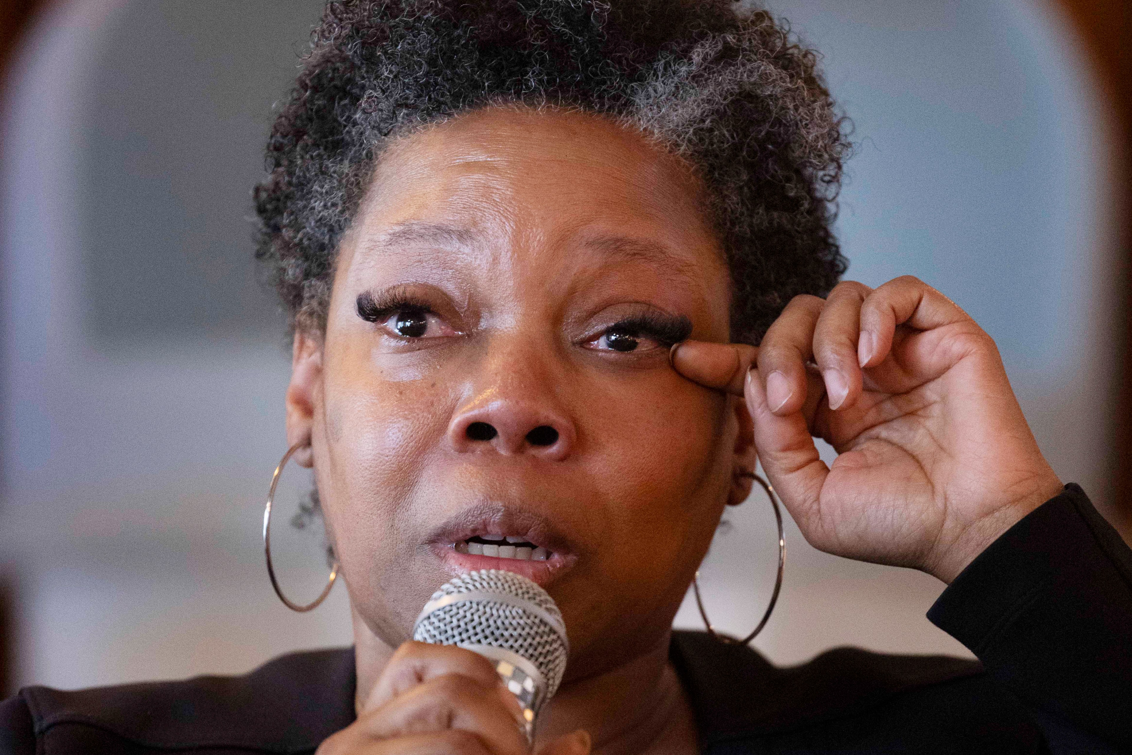 A person with short curly hair and hoop earrings holds a microphone while wiping their eye. The expression is emotional and focused.