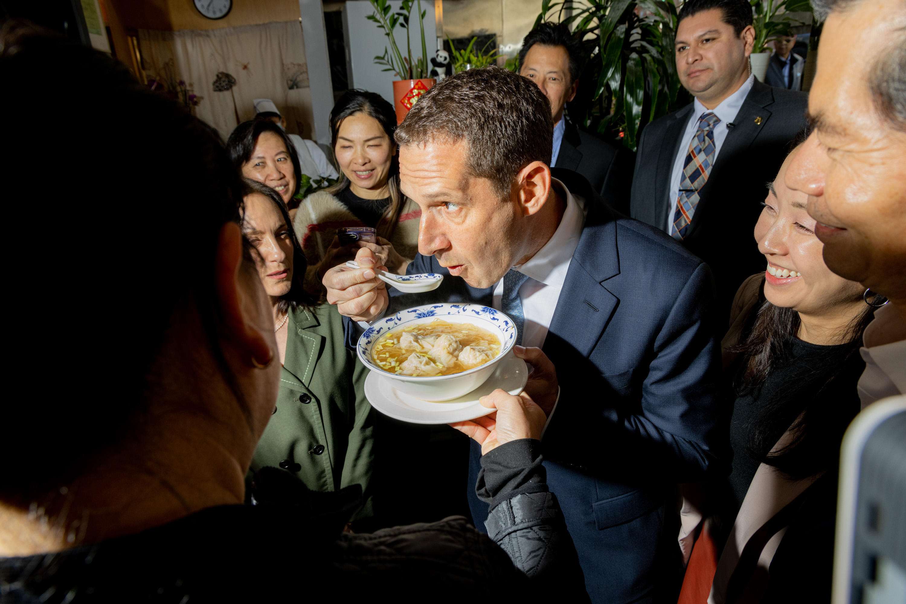 A man in a suit is sampling a bowl of soup with a spoon, surrounded by a group of smiling people in a warmly lit room with plants in the background.