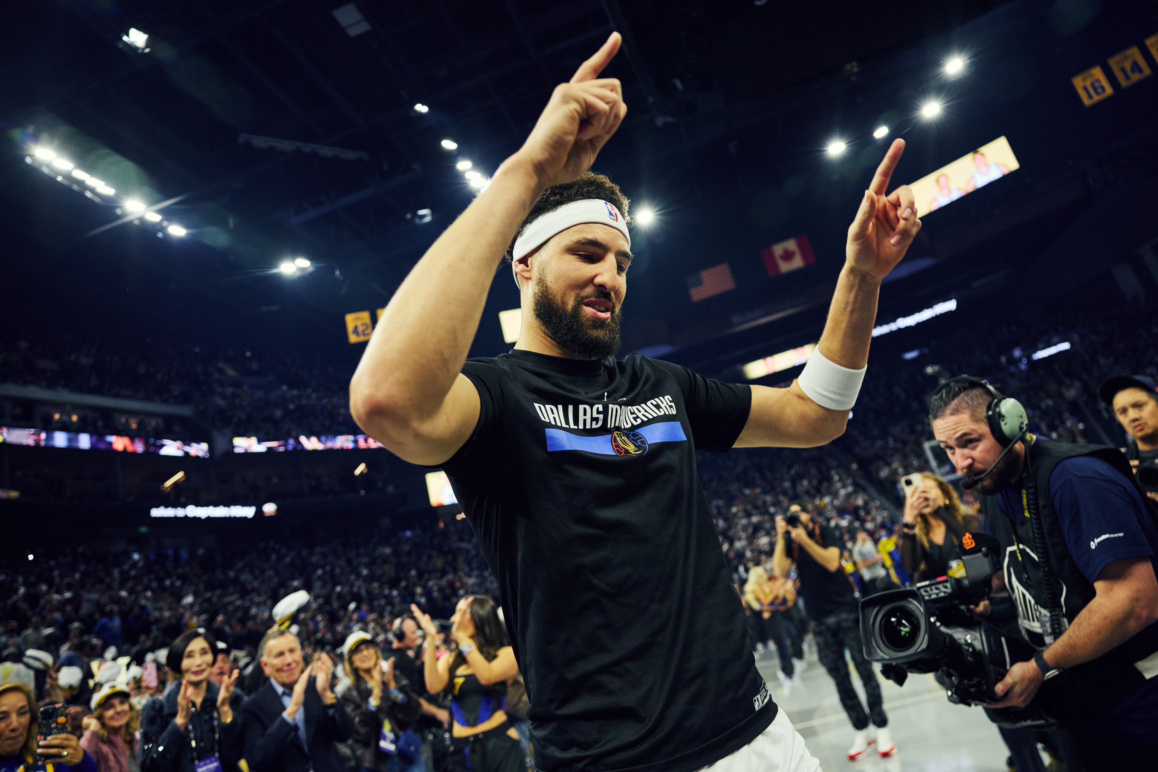 A basketball player, wearing a Dallas Mavericks shirt and headband, raises his hands in celebration on a well-lit court, surrounded by cheering fans and a cameraman.