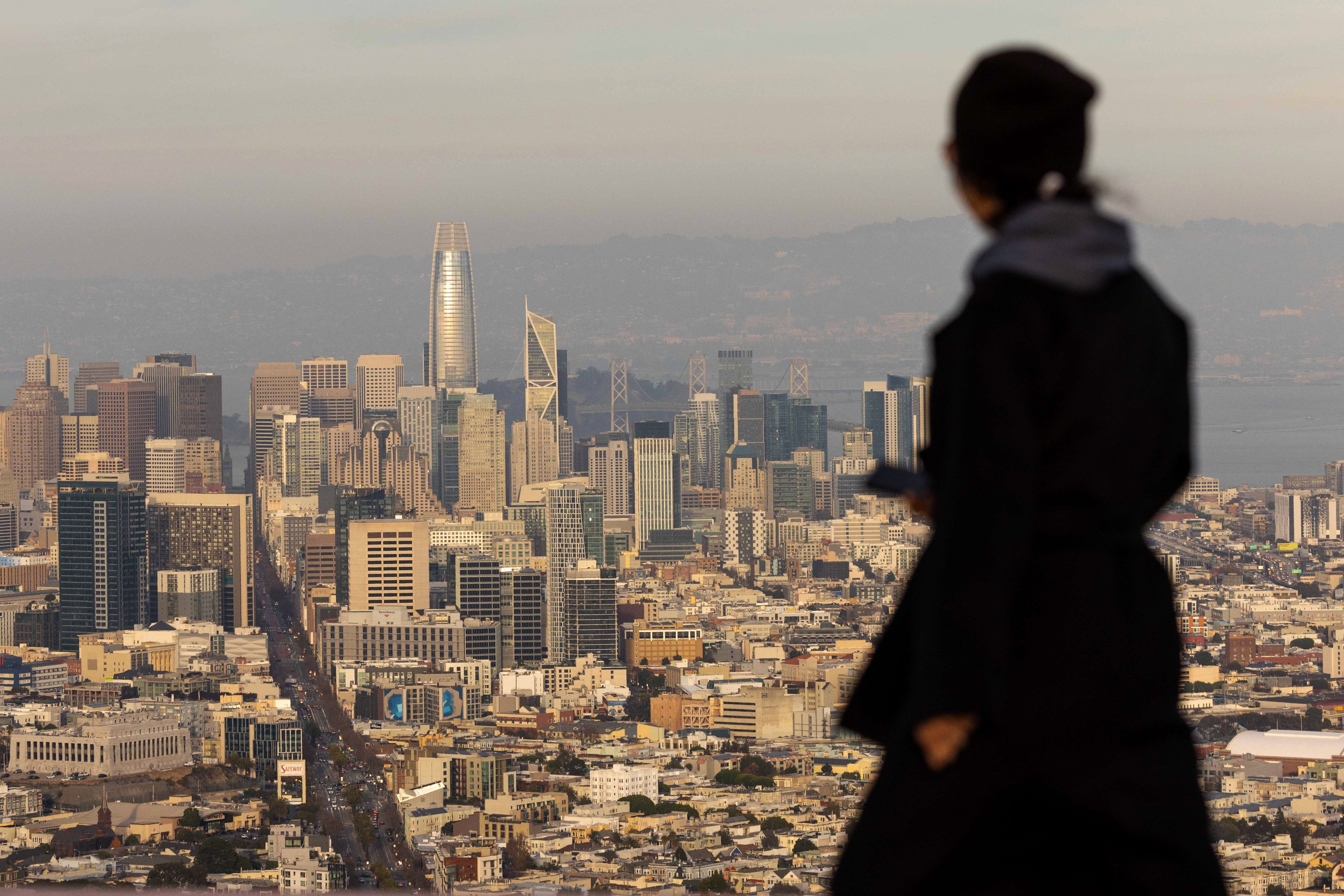 A person in silhouette stands overlooking a cityscape with tall skyscrapers and a hazy sky, creating a sense of depth and scale in an urban environment.