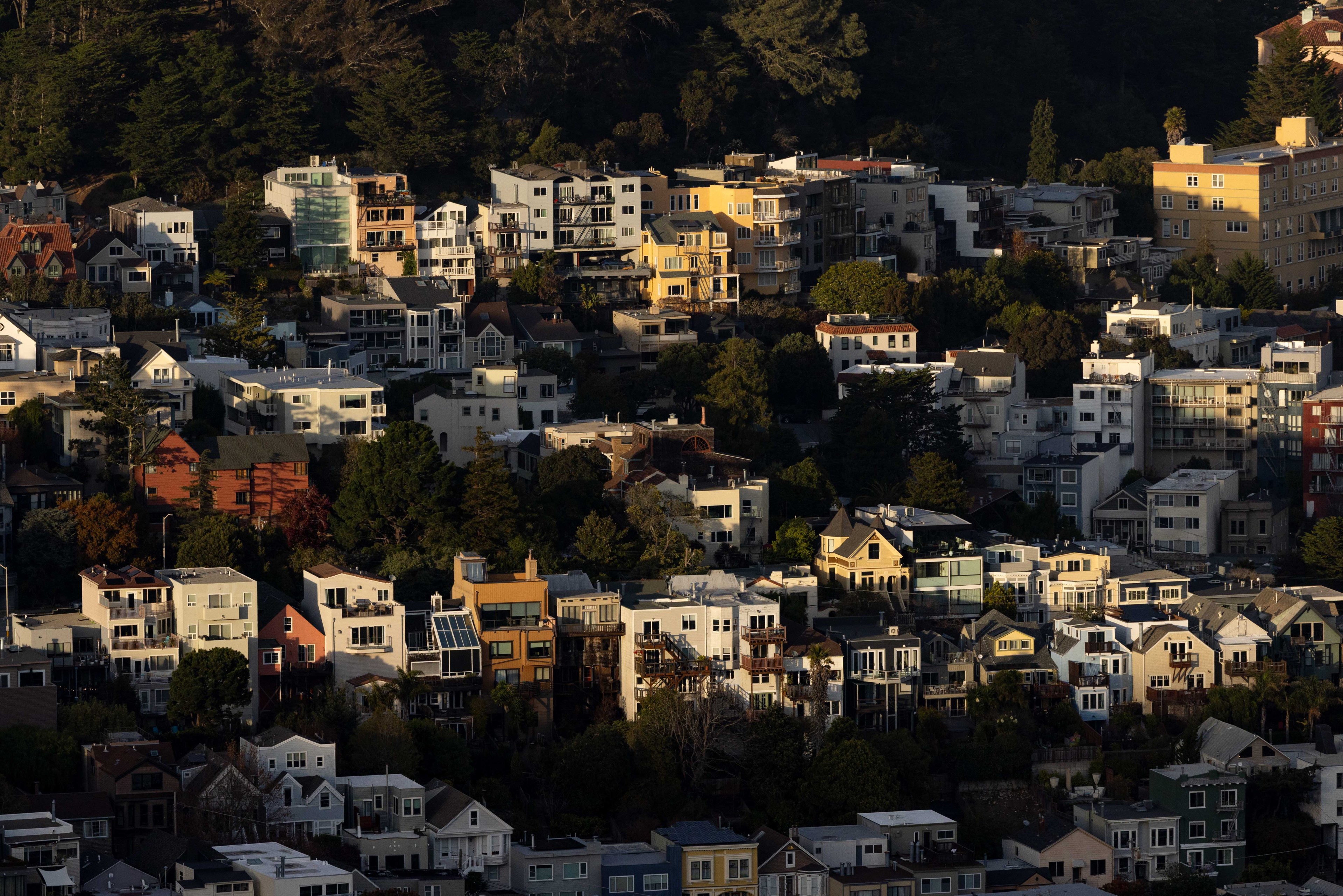 A densely packed hillside neighborhood with various styled houses surrounded by trees, bathed in warm sunlight, casting shadows and highlights across the scene.