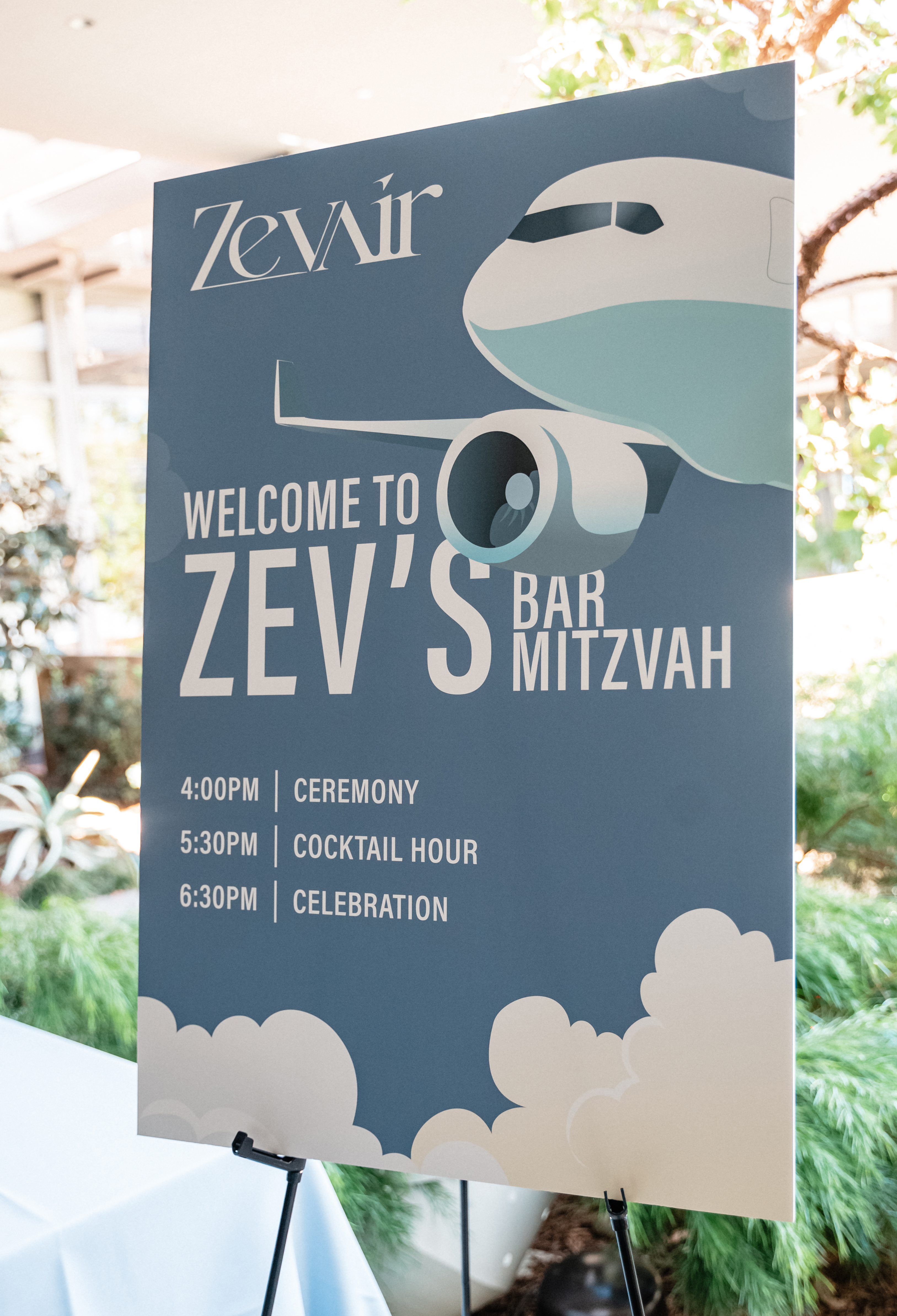 The image shows a sign with a blue background featuring an airplane illustration. It welcomes guests to a Bar Mitzvah with a schedule of events listed.