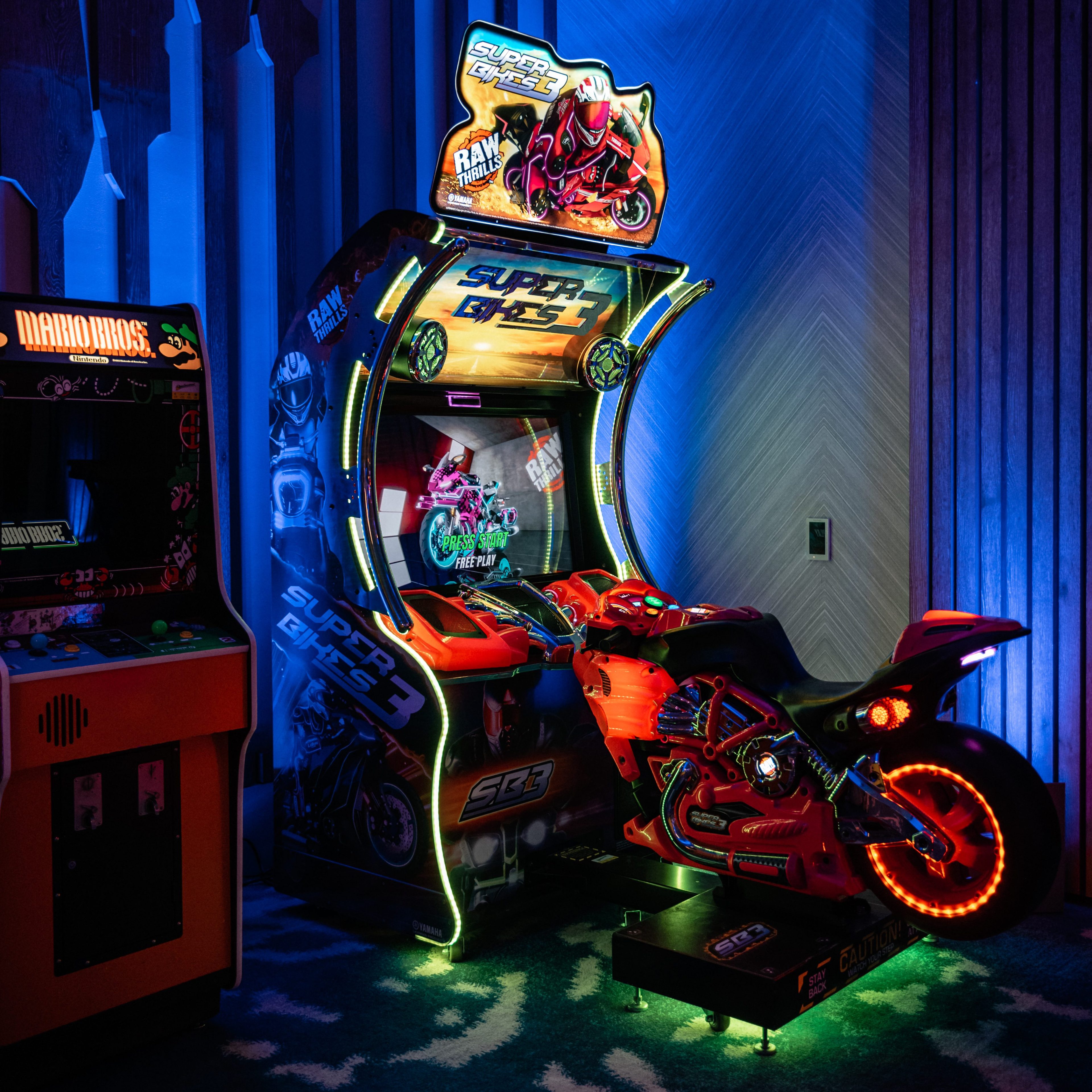 The image shows a brightly lit arcade room with a motorcycle racing game, &quot;Super Bikes 3,&quot; featuring a realistic red bike and vivid neon lights.